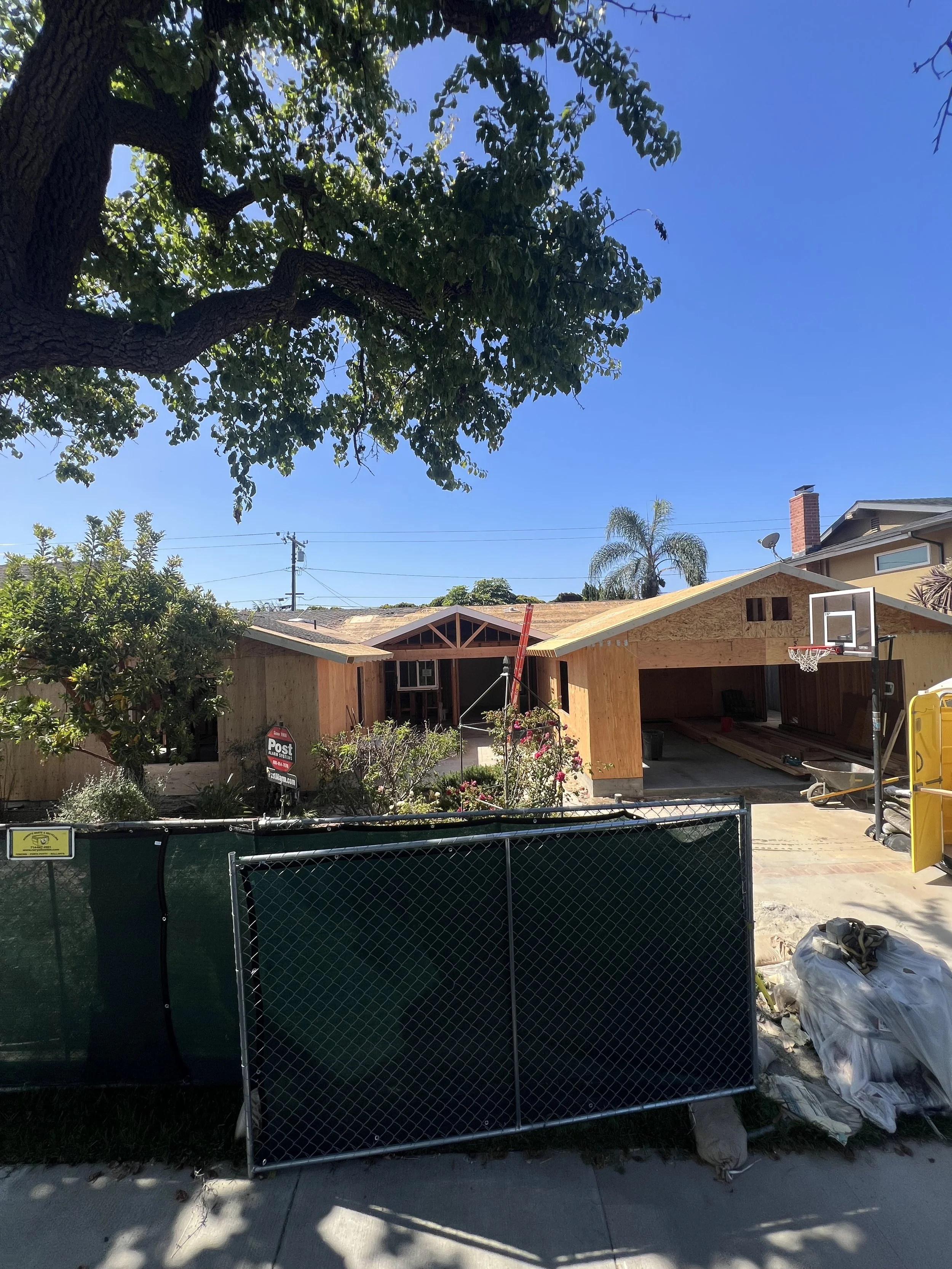 Construction site of a house with exposed wooden framing, a basketball hoop, and a tall tree with lush green leaves in the foreground.