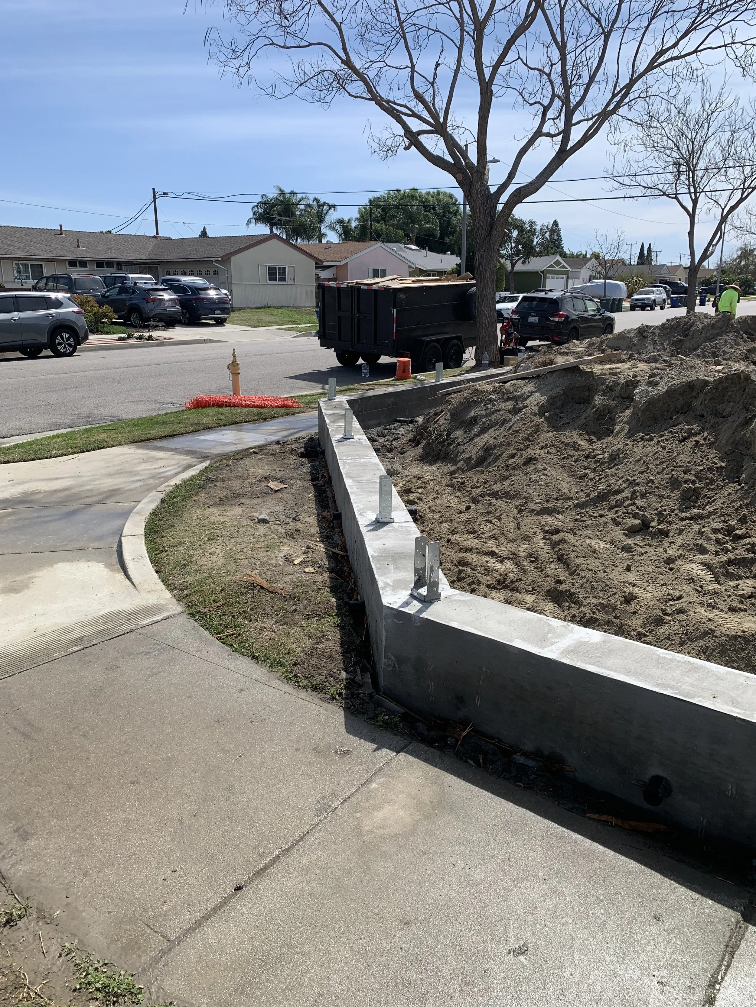 Sidewalk corner with ongoing construction, concrete curb and dirt mound, with parked cars, a black utility trailer, leafless trees, and houses in the background.