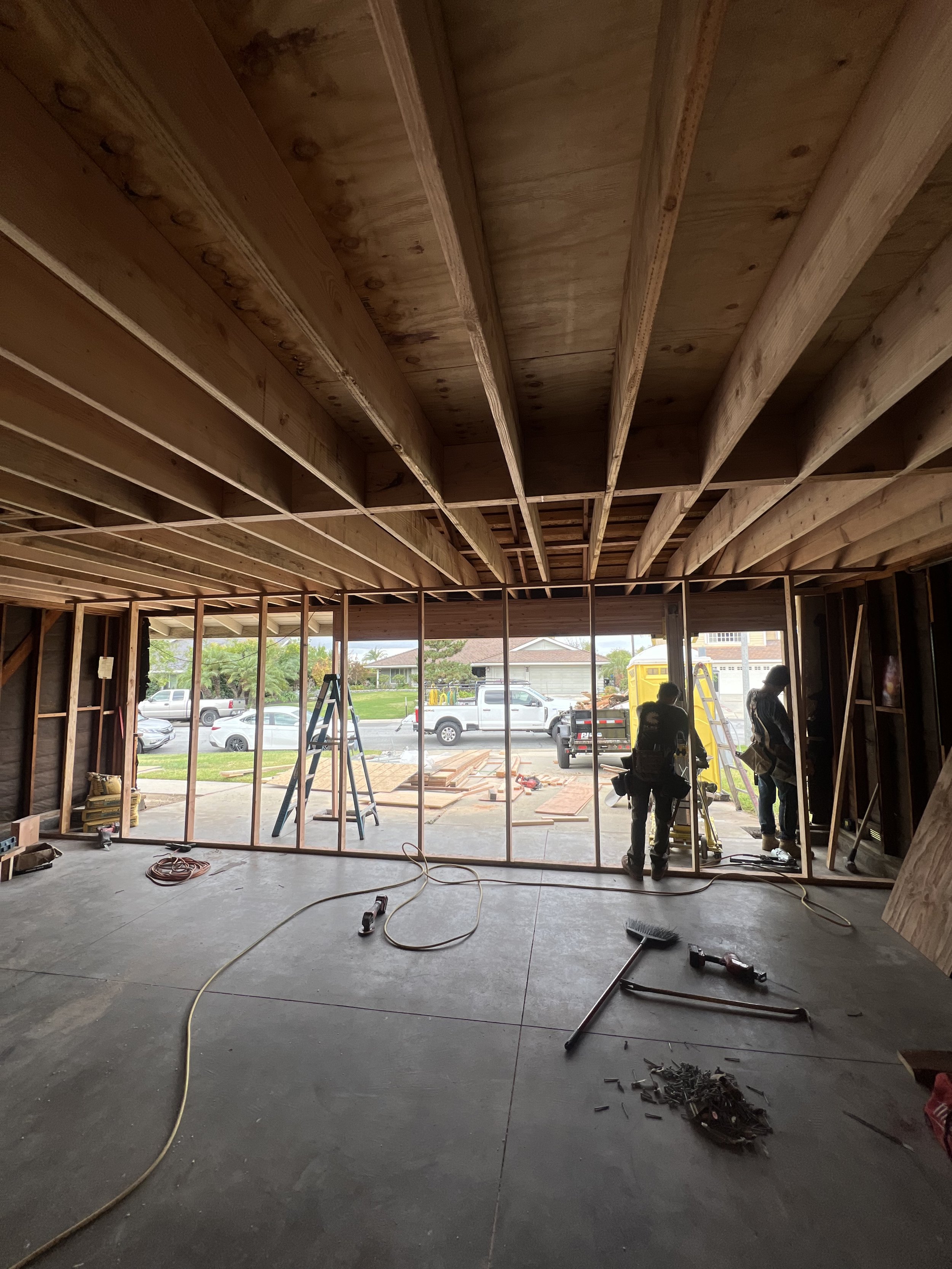 Construction workers framing the interior of a house, with viewed garage or front yard through large openings, construction tools and materials scattered on the floor.