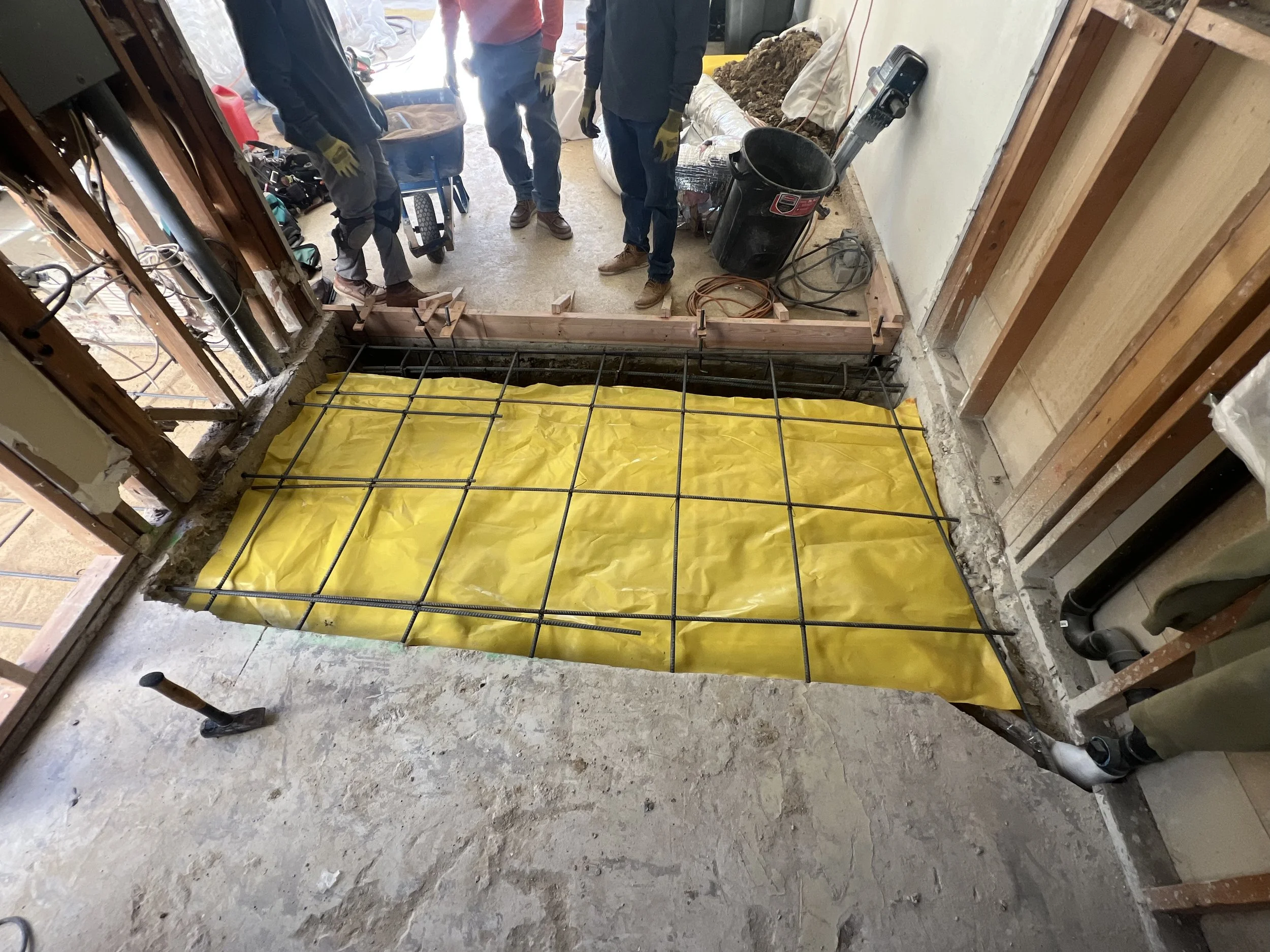 Construction workers install rebar, yellow waterproofing membrane, and formwork for a concrete floor or foundation in a residential renovation project.