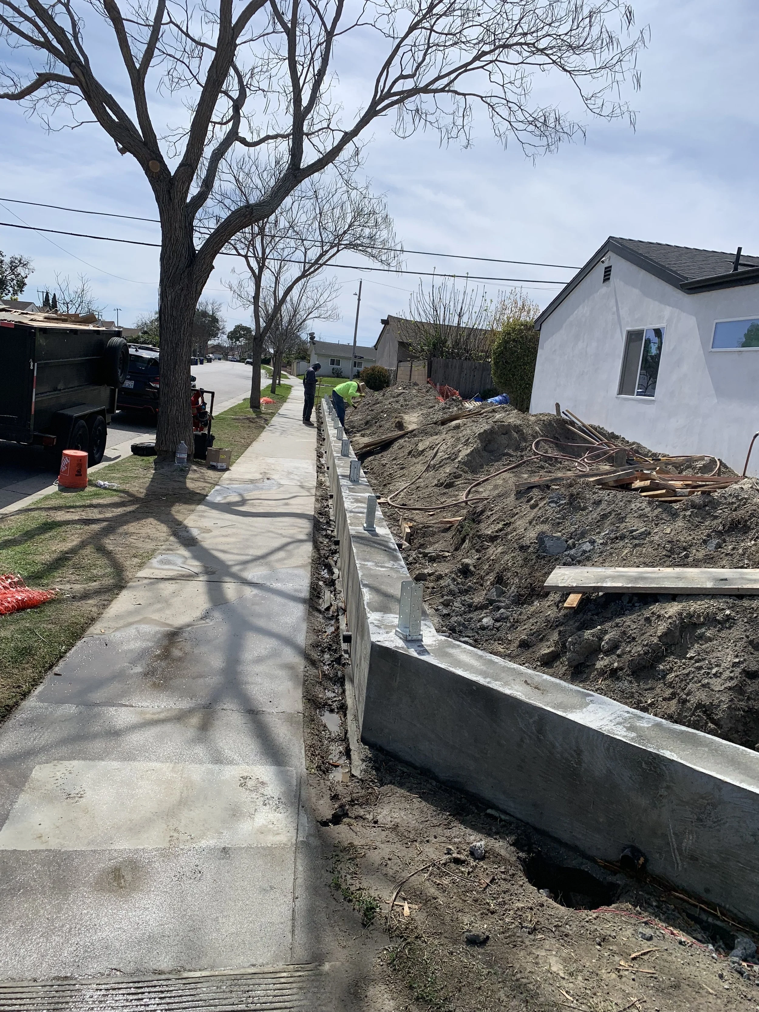 Construction workers working on sidewalk repair with a concrete barrier, dirt piles, and construction equipment on the side of a residential street with trees and houses.