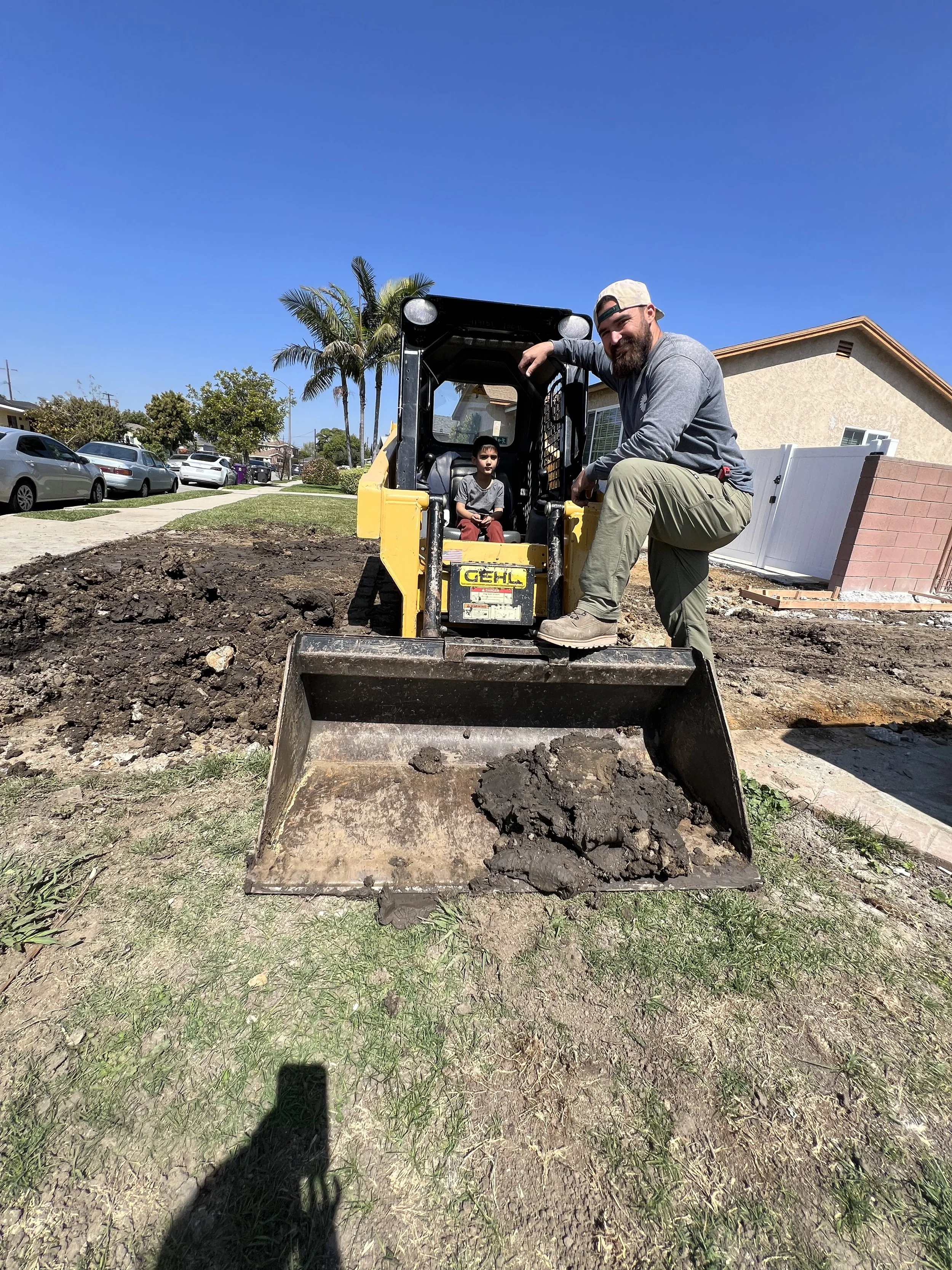 A man and a young boy on a construction site with a yellow skid-steer loader, dirt, and clear blue sky.