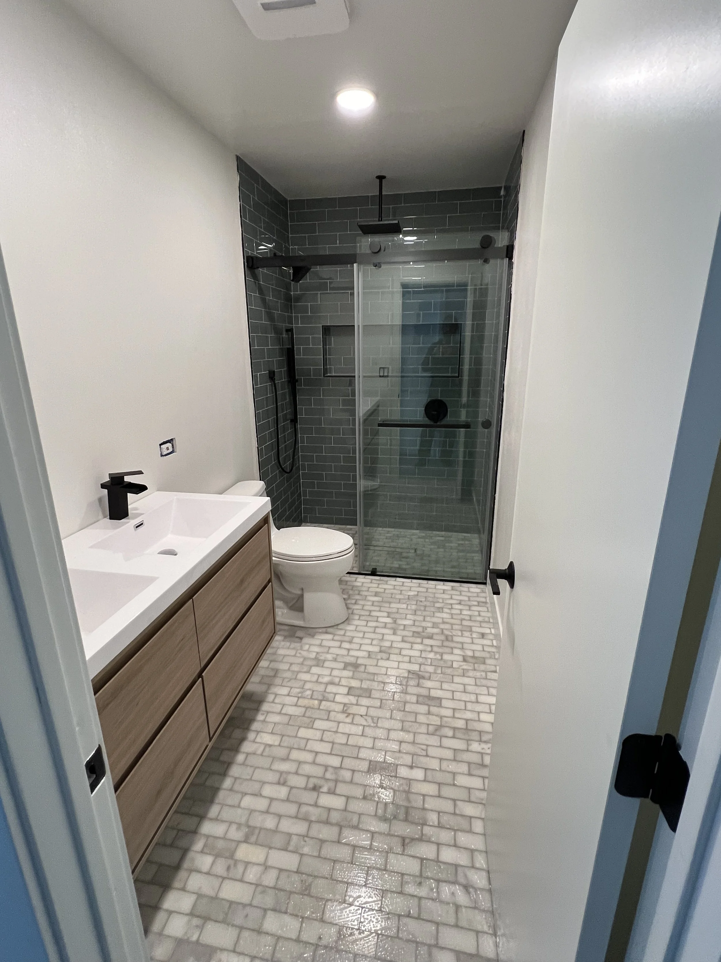 Modern bathroom with white walls, a wooden vanity with dual sinks, a black faucet, a white toilet, and a glass-enclosed shower with gray subway tiles and black fixtures.