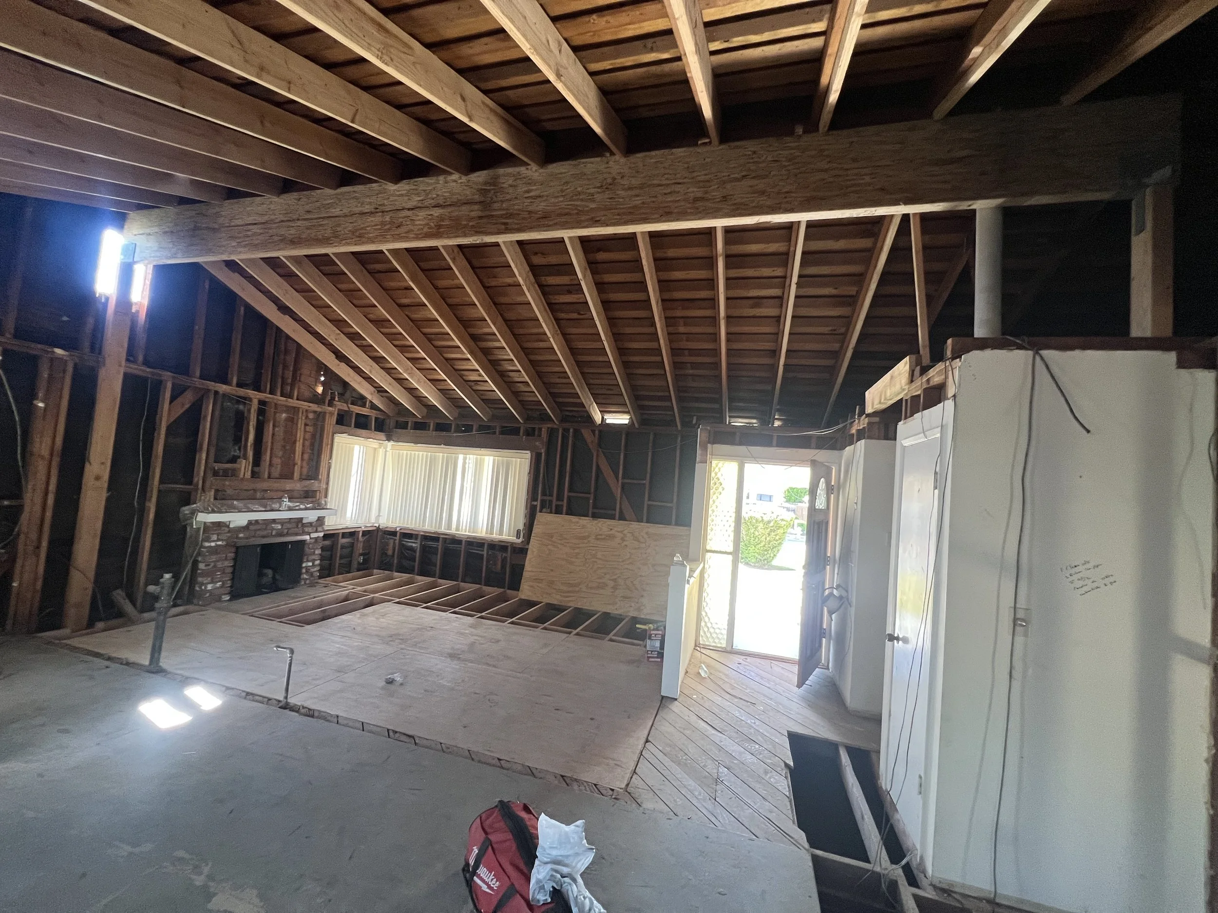 Interior of a house under renovation, with exposed wooden beams, partially finished flooring, a brick fireplace, and a small window near the ceiling, with construction materials and tools visible.
