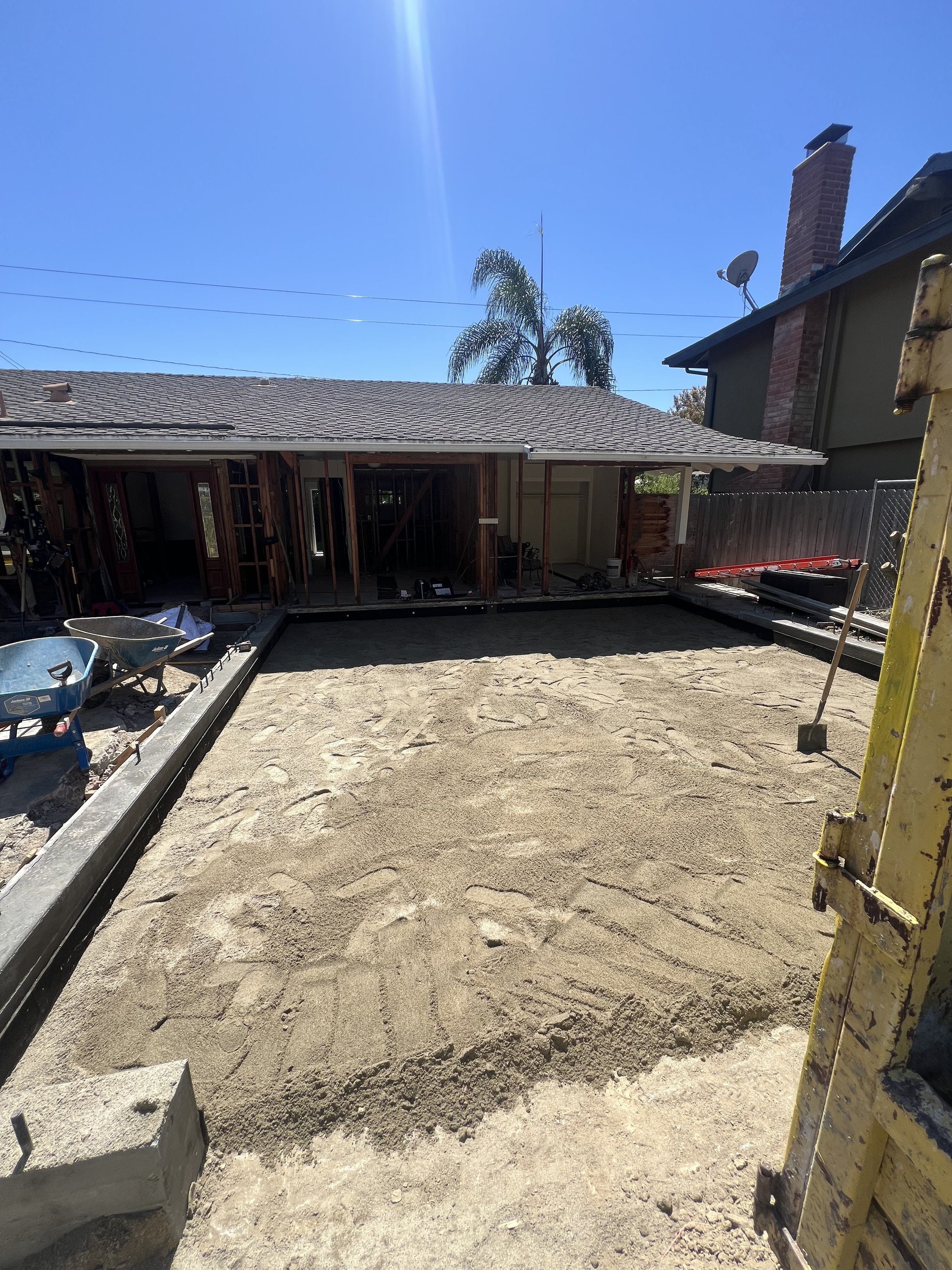 Construction site with a prepared level ground, wheelbarrows, and building materials, with a house under renovation in the background under a clear blue sky.