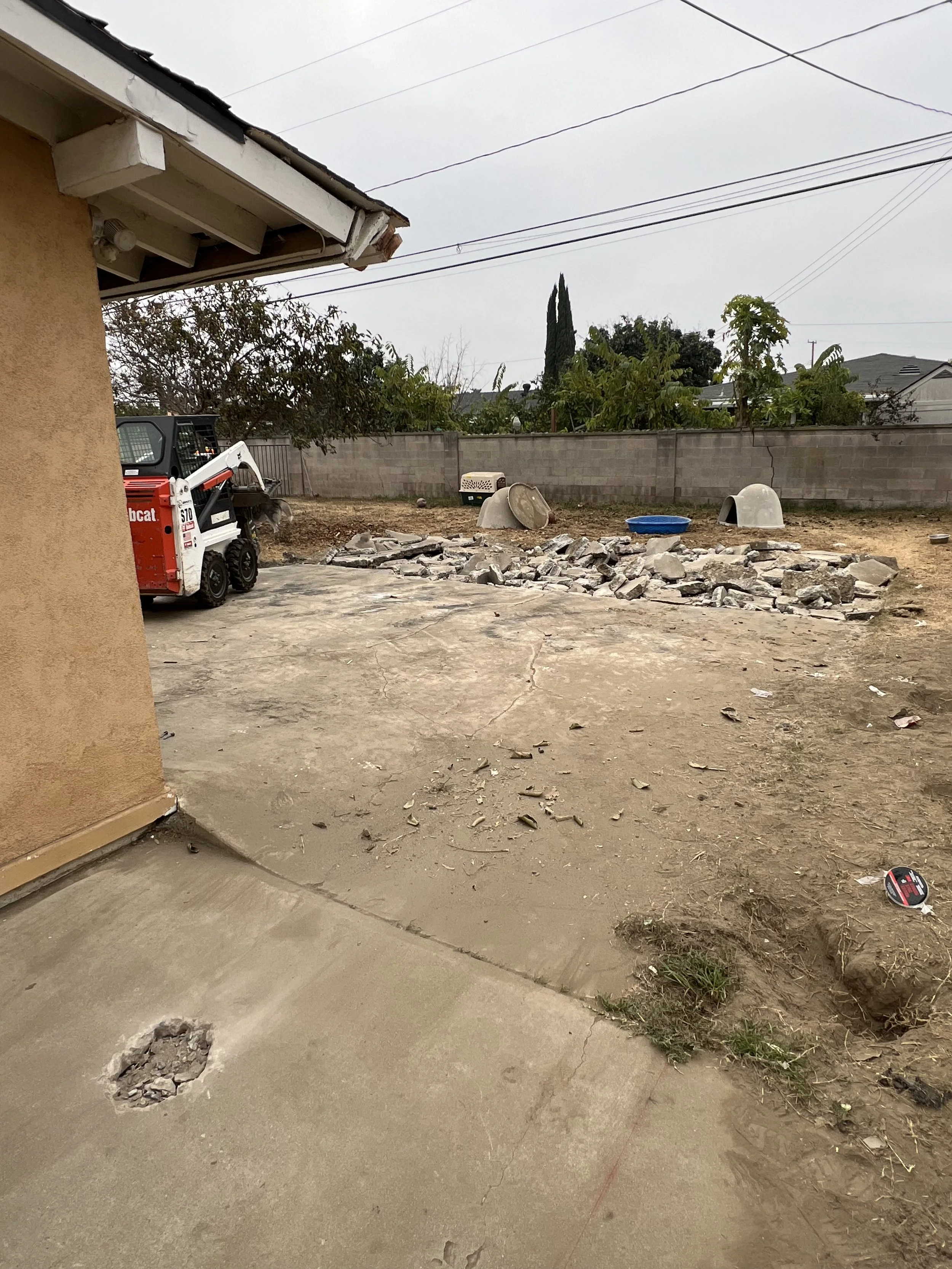 View of a backyard with a partially removed concrete slab, construction debris, a small skid steer, and a dog house, with a brick wall, trees, and cloudy sky in the background.