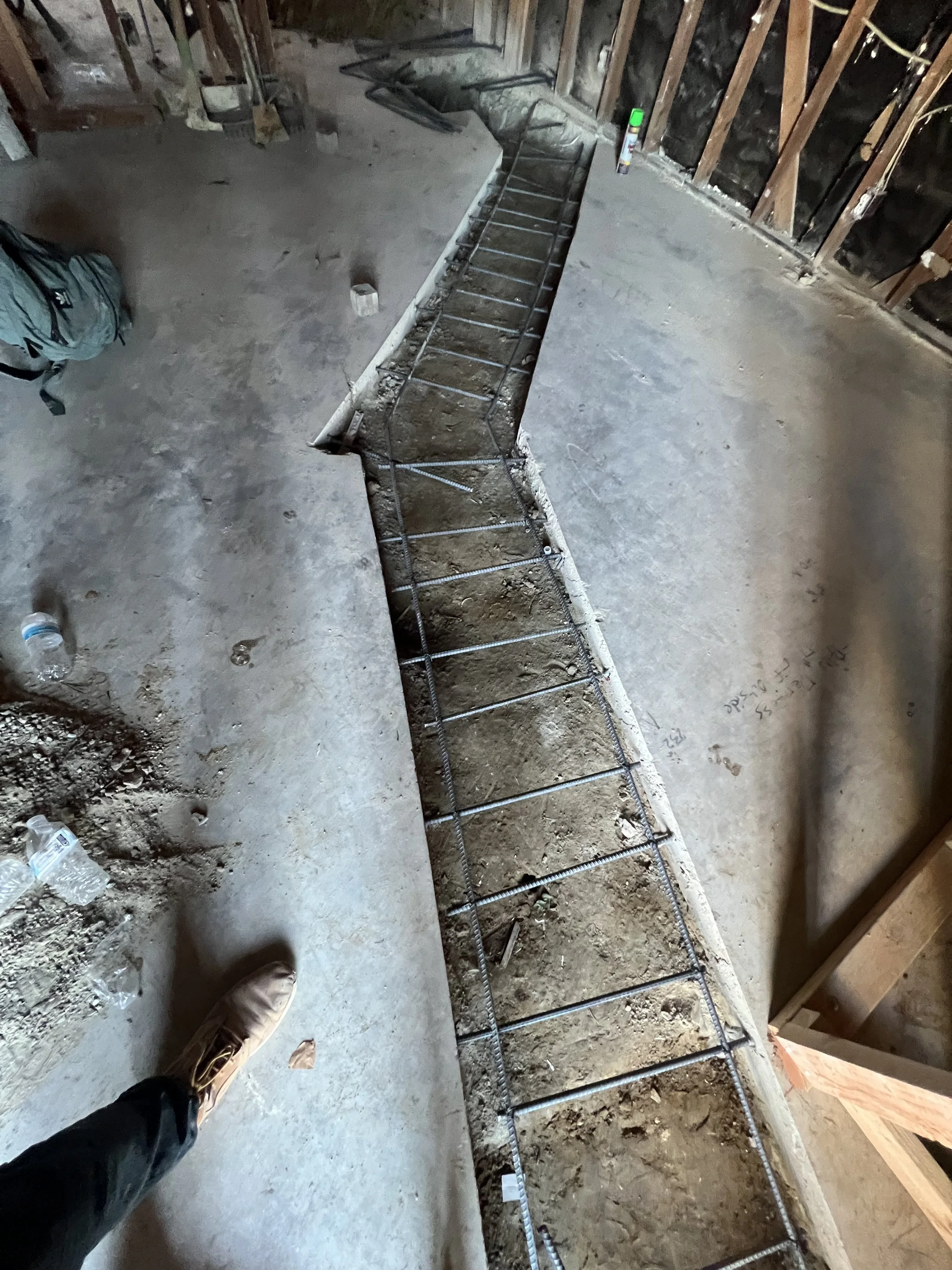 Construction site showing a trench with rebar reinforcement for a concrete pour, partially poured concrete around the trench, and various tools and supplies scattered around the area.