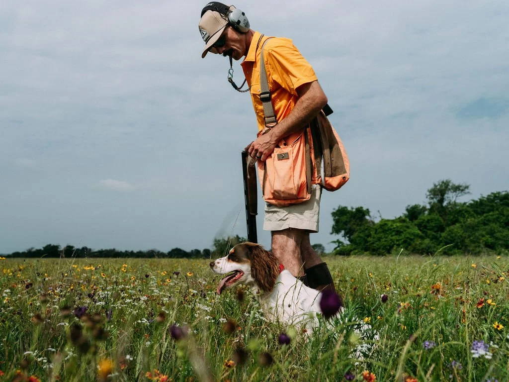 A man in a Poncho shirt, shorts, and a hat with a headset, holding a walking stick, standing in a field of wildflowers with an English Cocker sitting beside him.