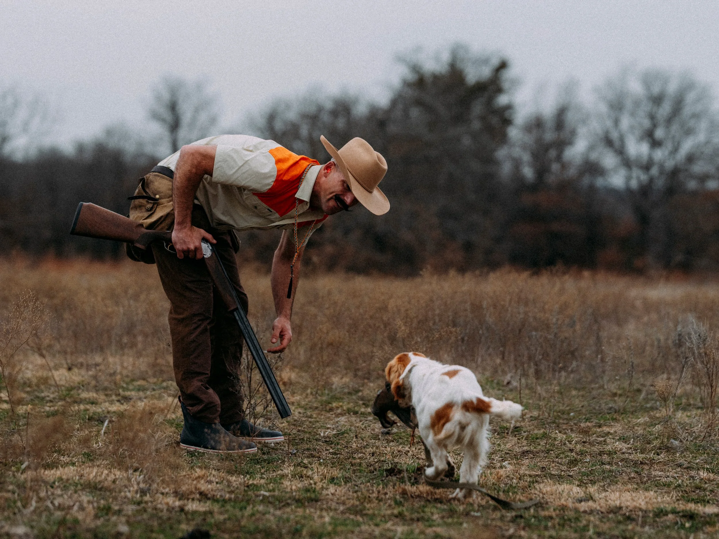 A man wearing a large tan cowboy hat, khaki shirt with orange accents, and brown pants, holding a shotgun, bends down to pet an English Cocker Spaniel in an open field with dry grass and a background of leafless trees.