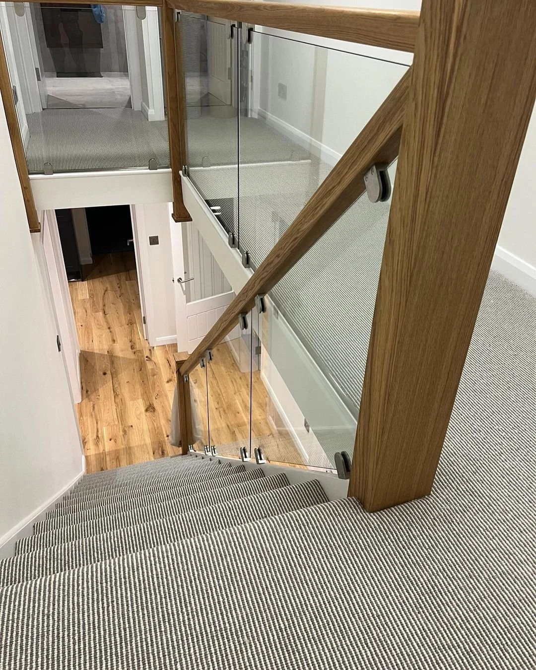 View of a staircase with a glass railing and wooden handrail, leading down to a hallway with wooden flooring and white walls, in a modern home.