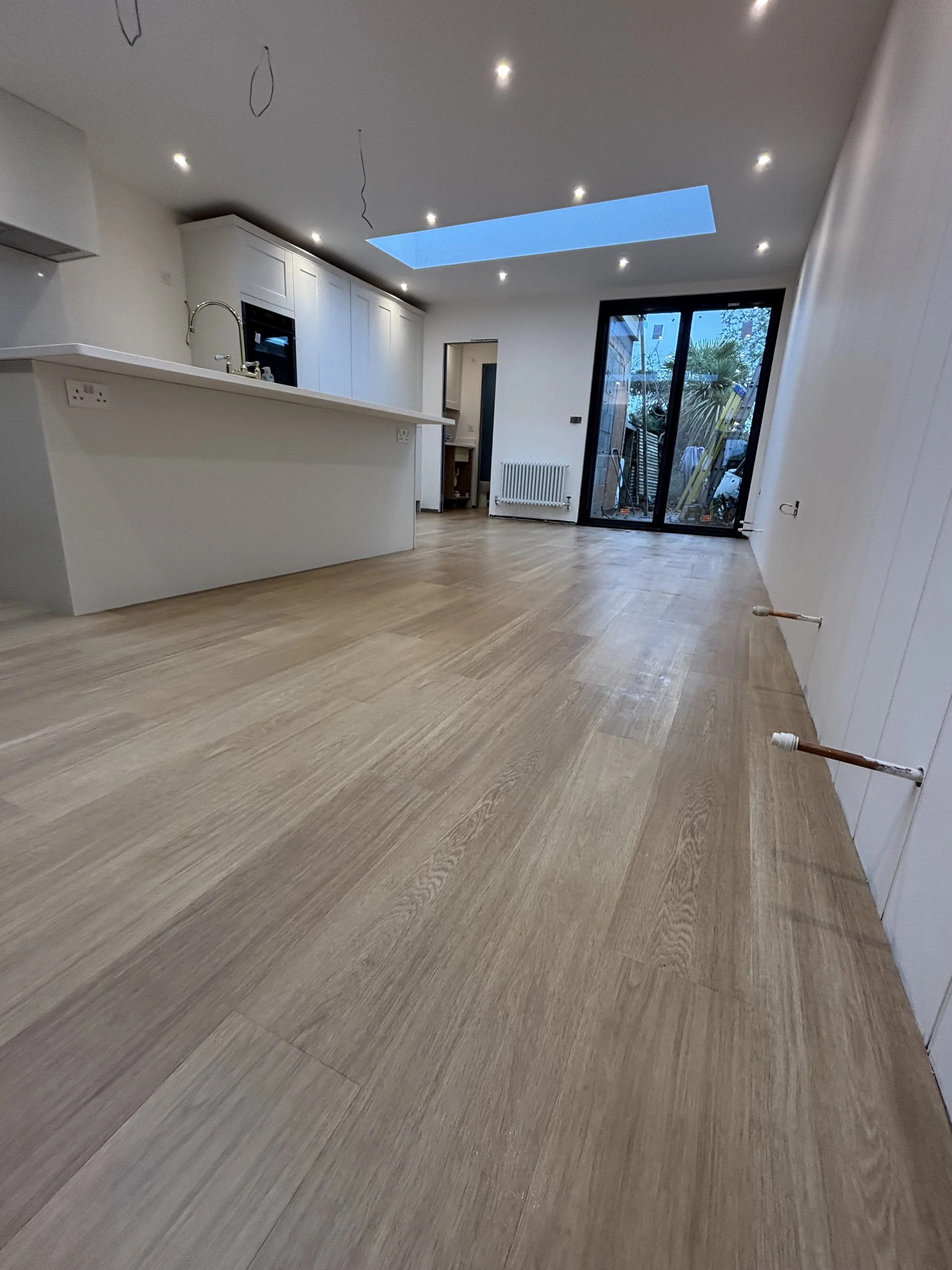 Empty modern kitchen with white cabinets, a kitchen island, wooden flooring, and large glass doors leading outside. The ceiling has recessed lights and a skylight.