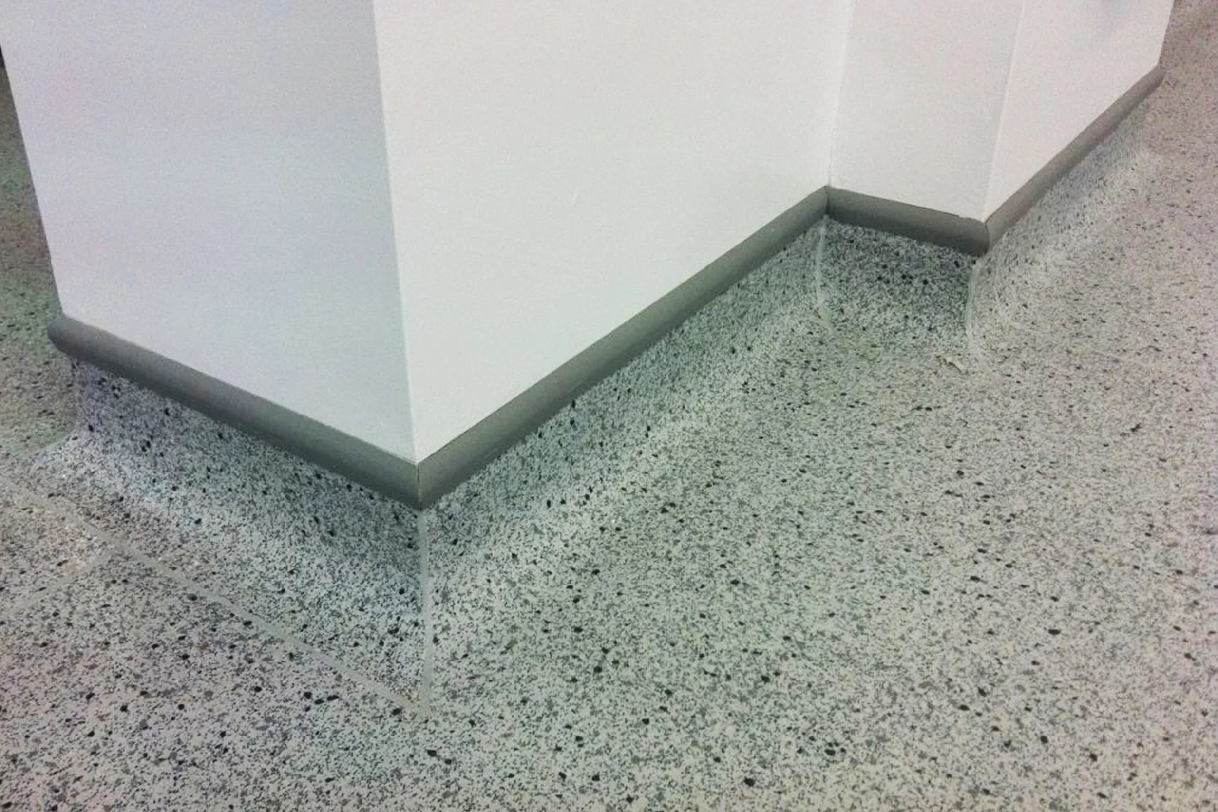 Close-up of a corner in a building showing the transition between a speckled terrazzo floor and a white painted wall with gray baseboard molding.