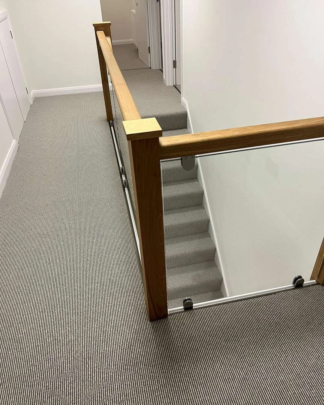 Image of a staircase leading to a lower level with a wooden handrail, surrounded by a safety gate, in a residential corridor with white walls and striped carpet.