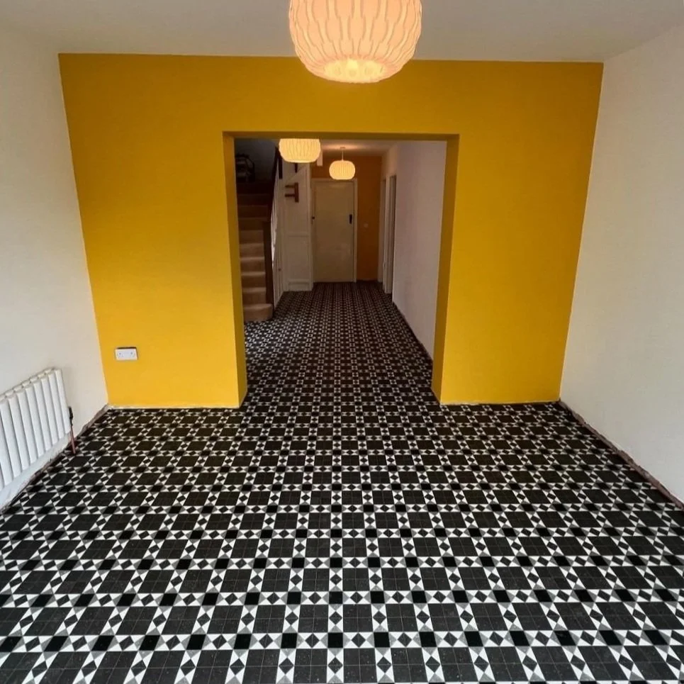 Interior view of a house with black and white patterned tile flooring, white walls, a yellow accent wall framing a doorway, and hanging paper lantern ceiling lights.