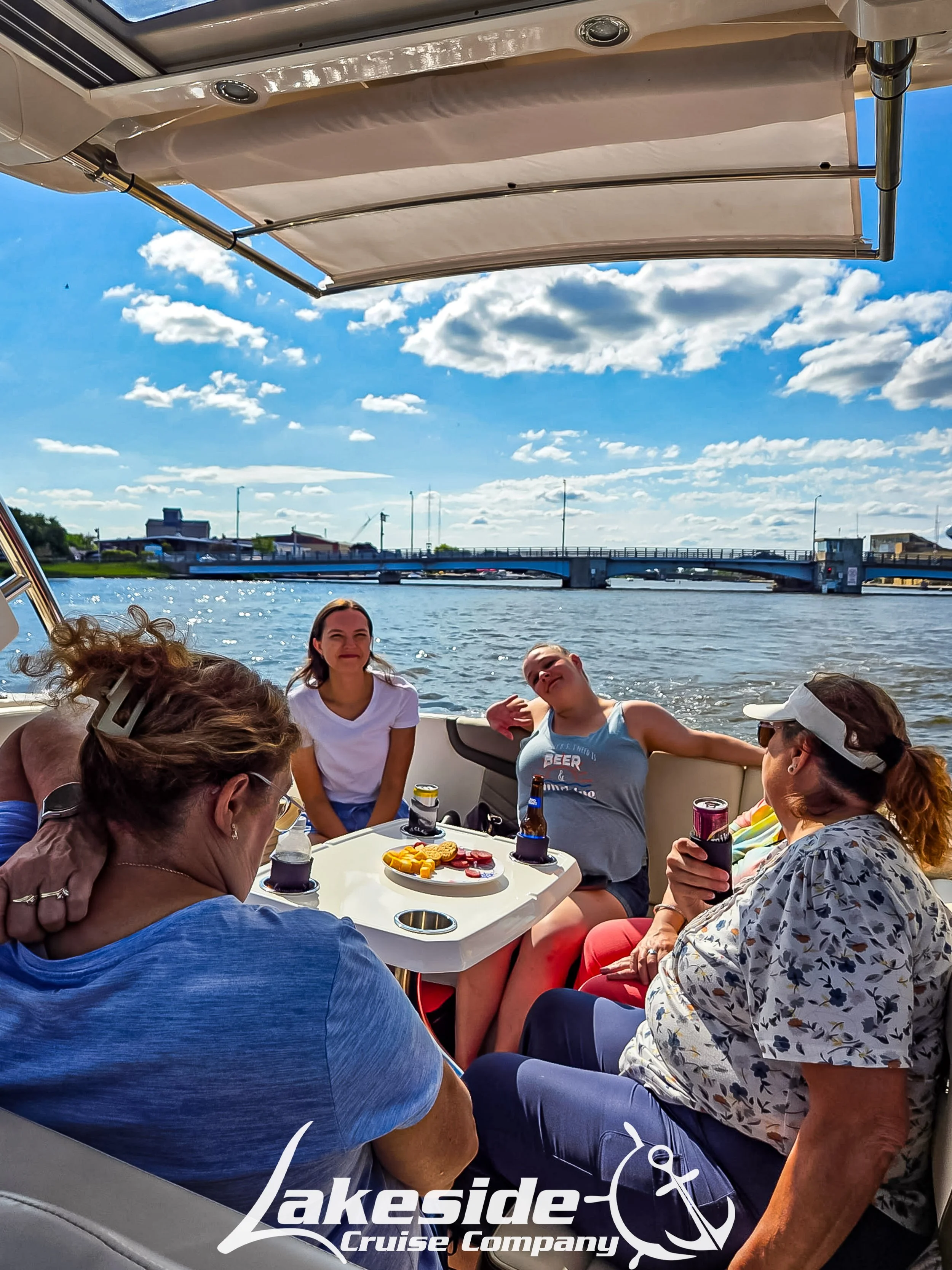 Group of people relaxing and enjoying drinks on a boat with water and bridge in the background, under a partly cloudy sky, with Lakeside Cruise Company logo in the corner.