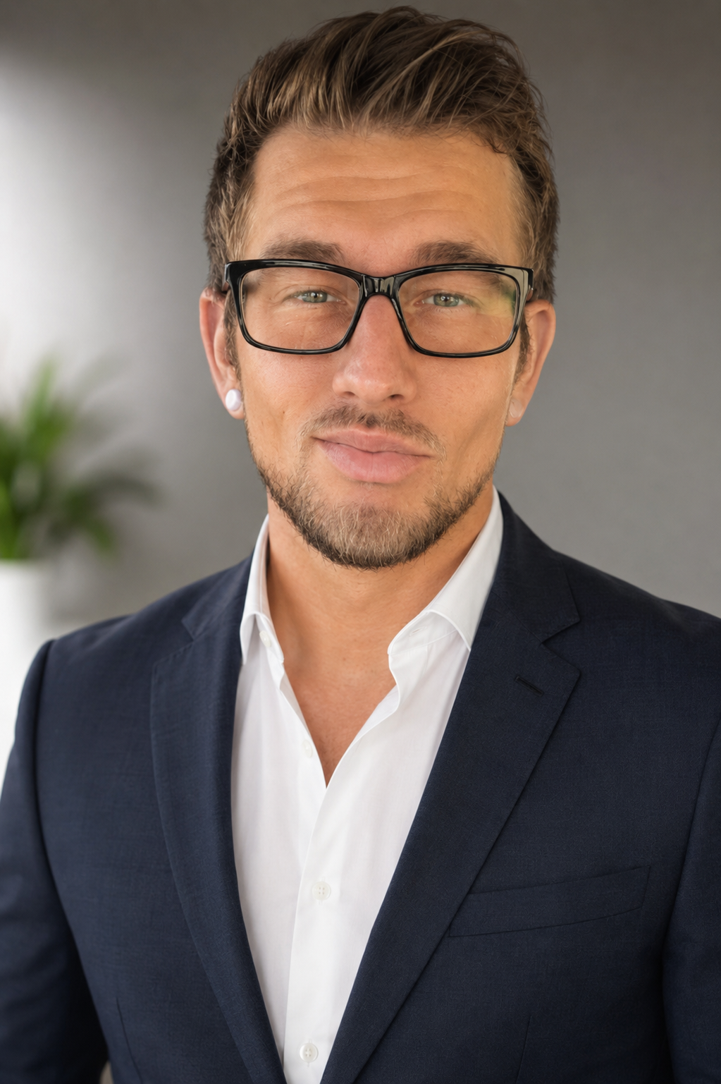 A young man with glasses, earrings, and a necklace taking a selfie indoors with a blurred background of household items and plants.