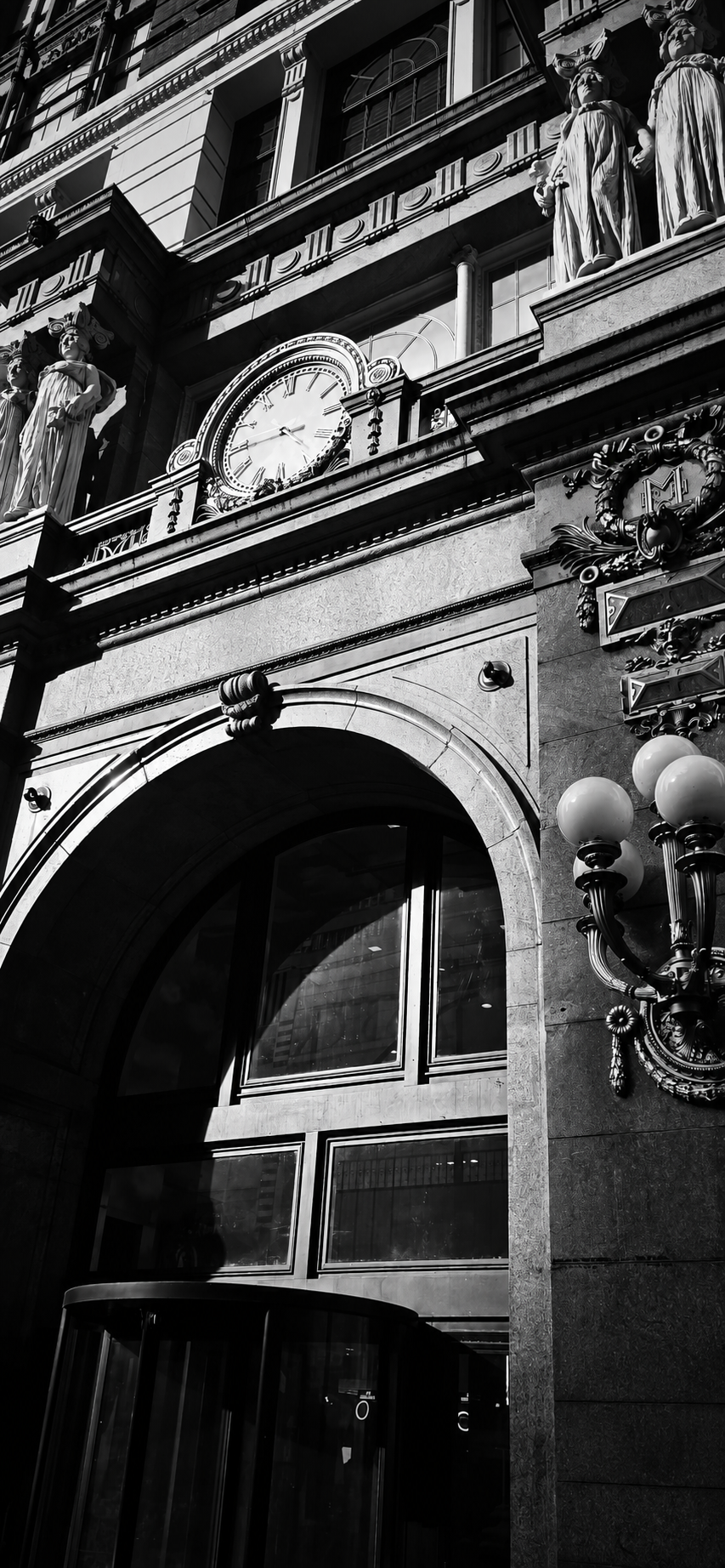 Black-and-white view of an ornate historic building facade with a large clock, statues, carved decorations, arched entrance, and globe lamps