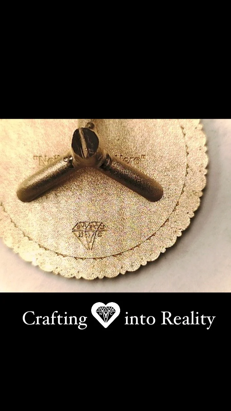 Vintage bronze necklace pendant with two crossed bars on a scalloped, engraved paper card, above the text “Crafting into Reality.”