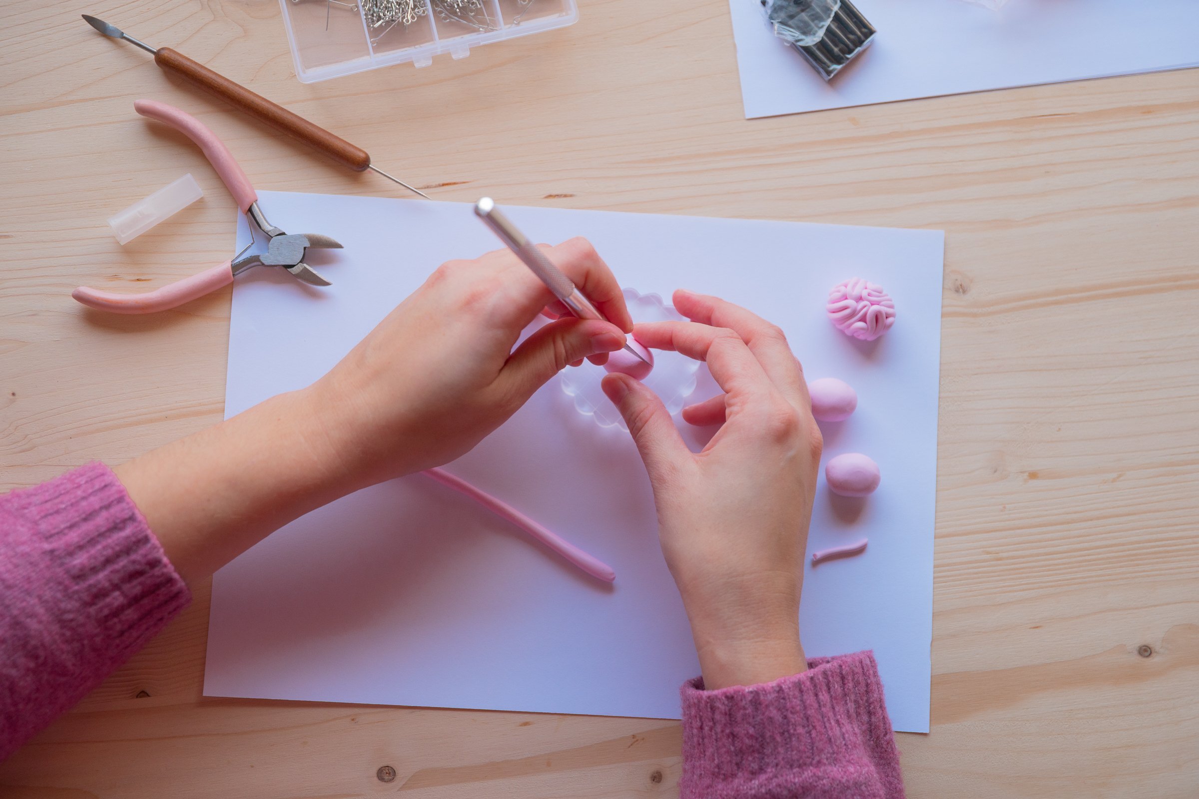 Person modelliert mit rosa Knete auf einem weißen Papier, umgeben von Knetwerkzeugen und Knetteilen in rosa auf einem Holzarbeitsplatz.