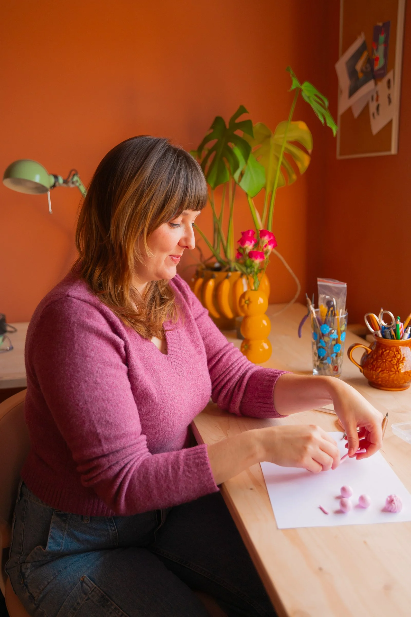Frau sitzt an einem Holztisch, arbeitet mit rosa Modelliermasse, im Hintergrund orange Wand, Pflanzen und Dekorationsgegenstände.
