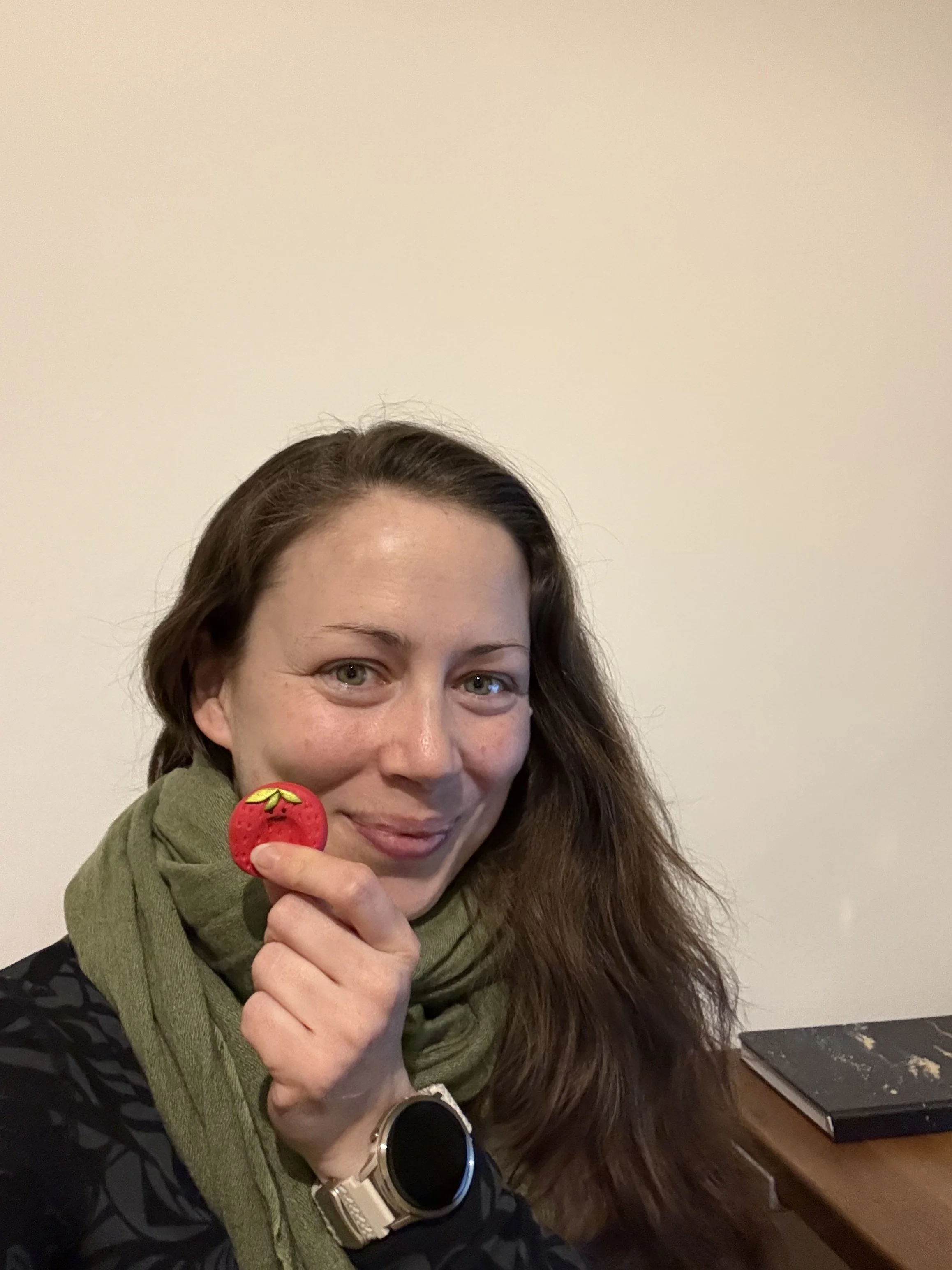 Frau mit grüner Halskette hält einen roten, strawberry-Formen Seifenblasen-Spielzeug vor Gesicht, sitzt an einem Tisch mit einem schwarzen Buch.