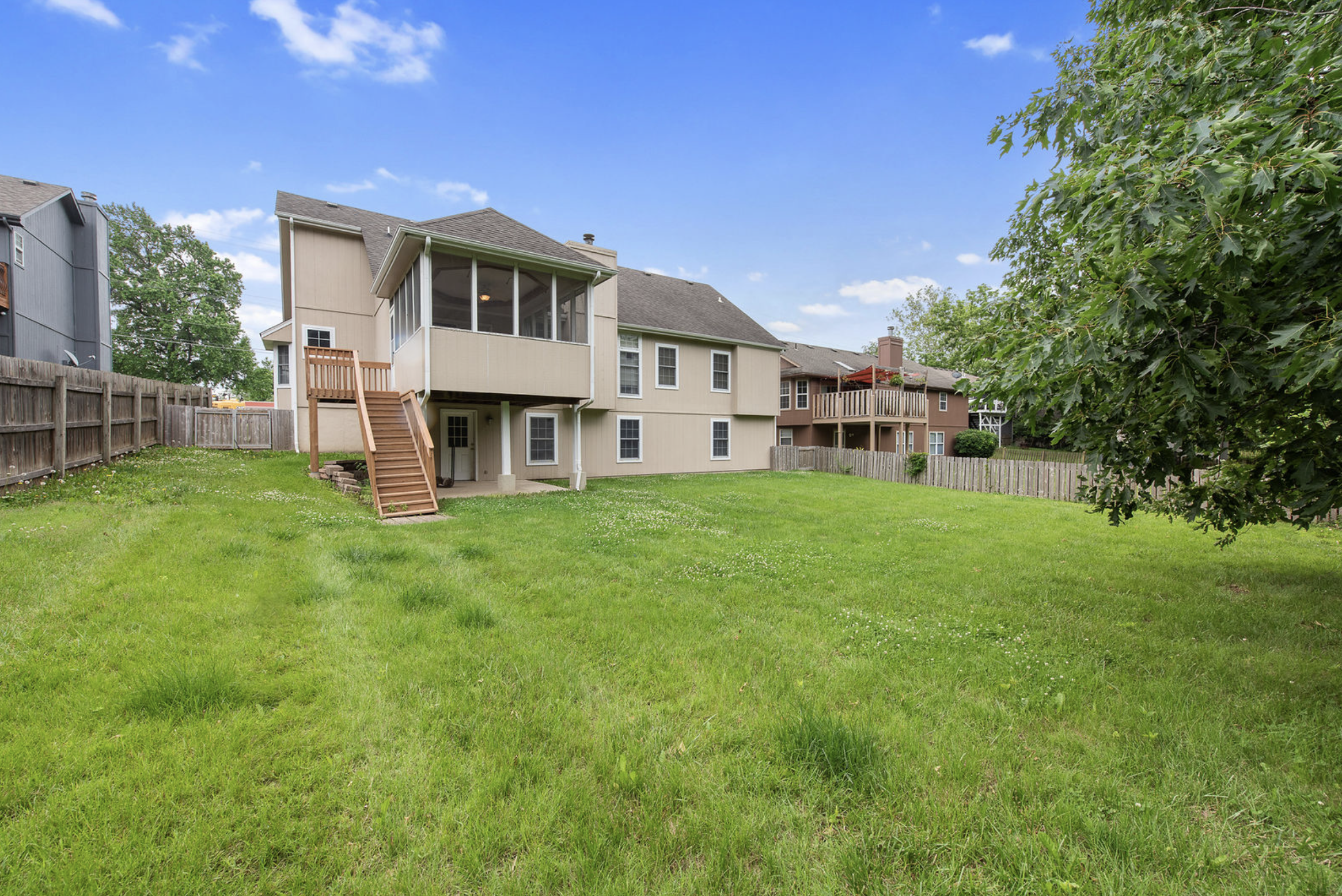 Backyard with green grass, fenced in on three sides, and a beige two-story house with a screened-in porch and an outdoor staircase leading to the ground.