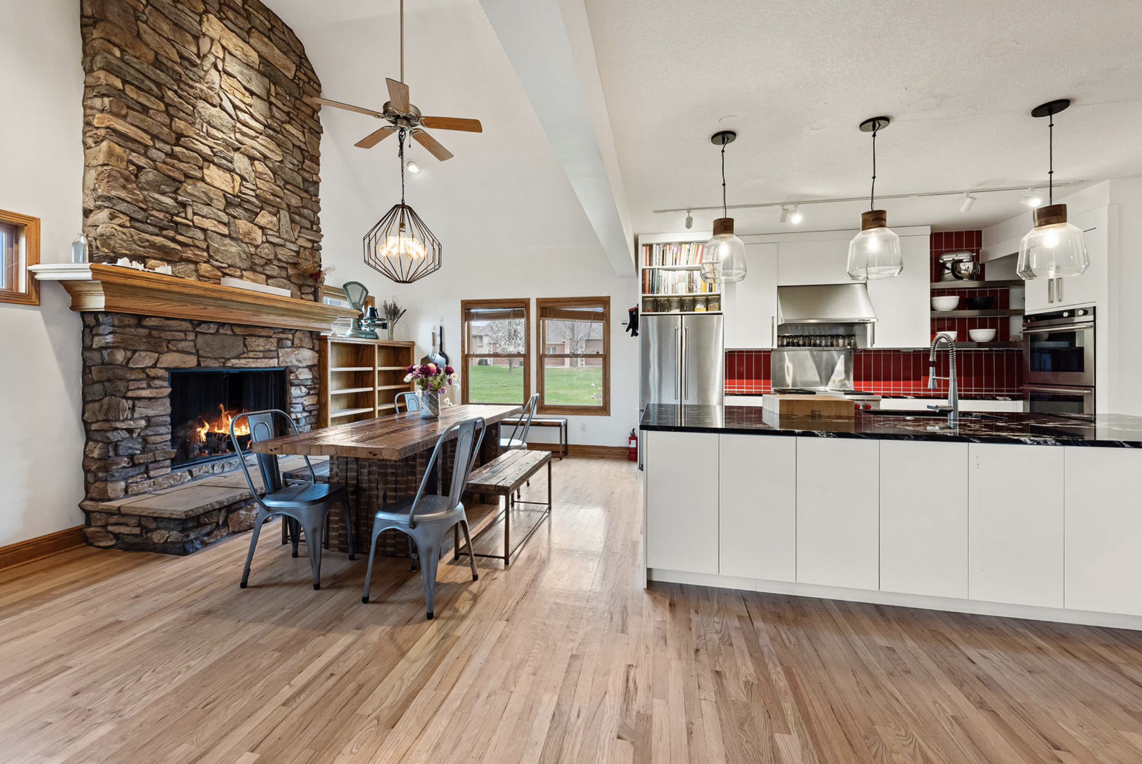 Open-concept kitchen and dining area with a stone fireplace, wooden dining table with chairs, and modern white kitchen with black countertops, pendant lights, and red tile backsplash.