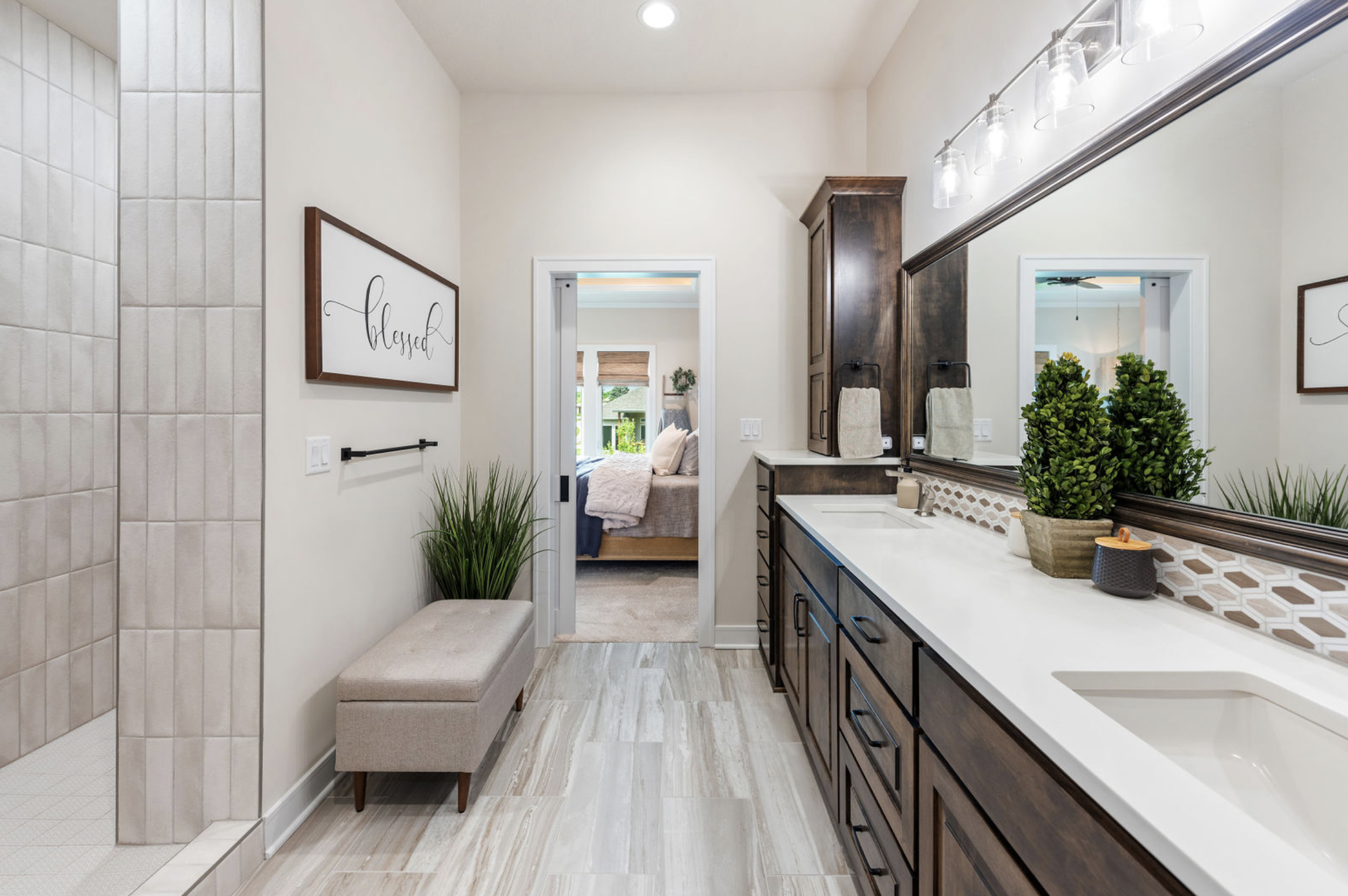 Bathroom with dual sinks, wooden cabinets, large mirror, plants, wall art, and a view into a bedroom.