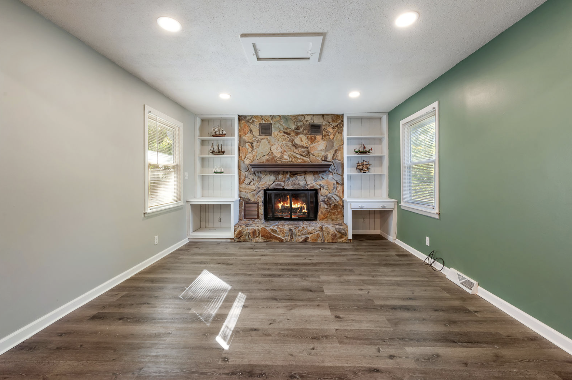 Empty living room with wood flooring, stone fireplace, built-in white shelves, and windows with blinds.
