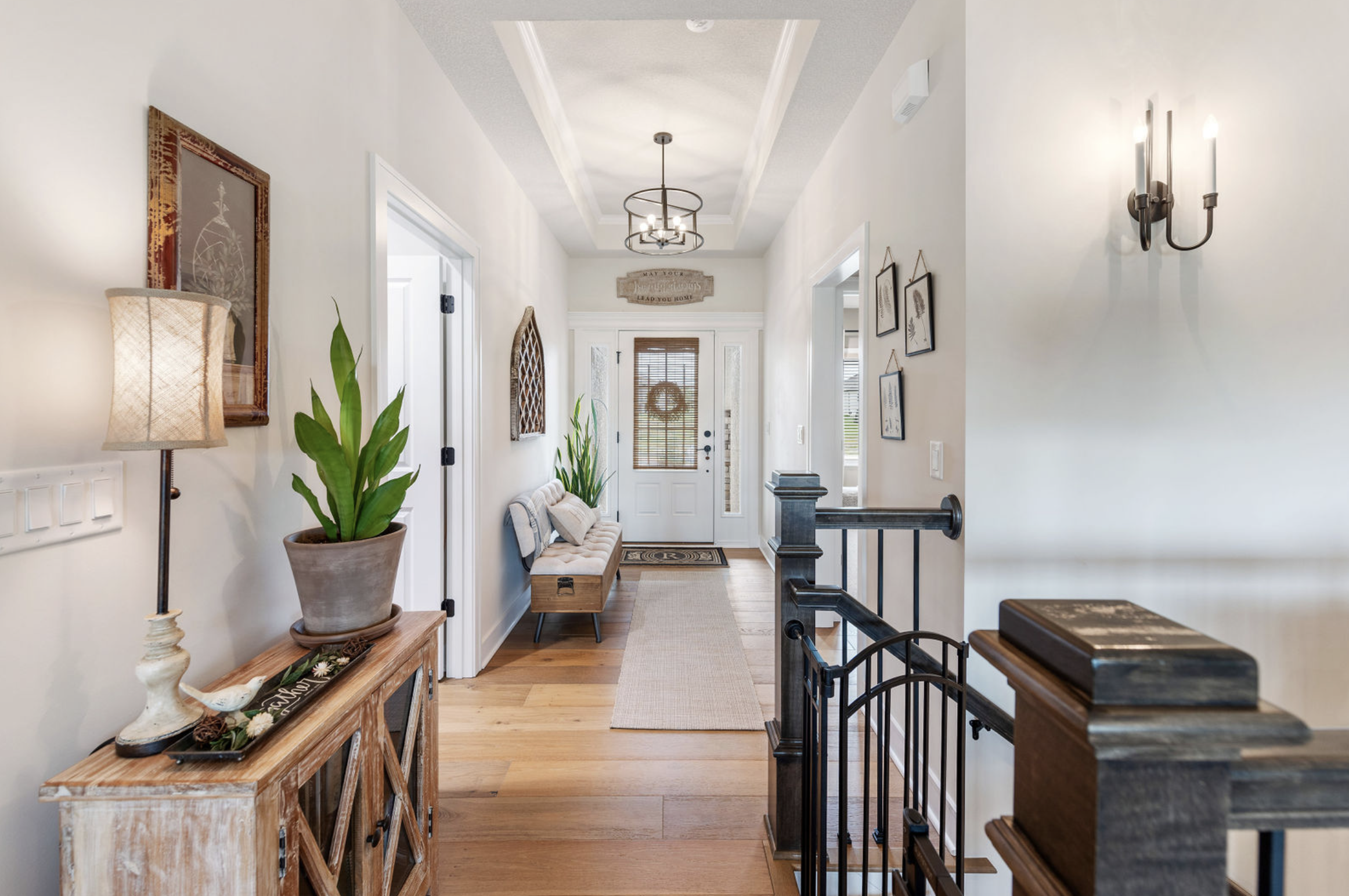 Entry hallway with white walls, wooden flooring, decorative wall art, a bench with cushions near the front door, potted plants, a wooden console table, and wall-mounted light fixtures.