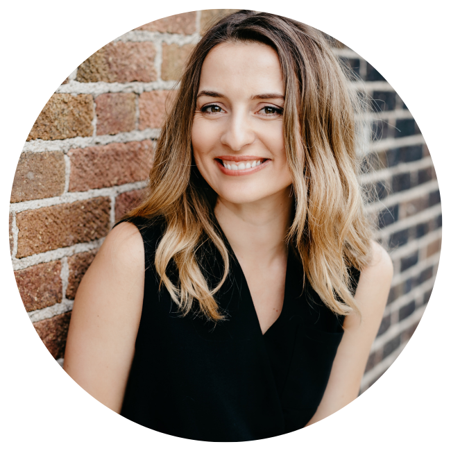 A young woman with shoulder-length wavy blonde hair smiling while leaning against a brick wall.