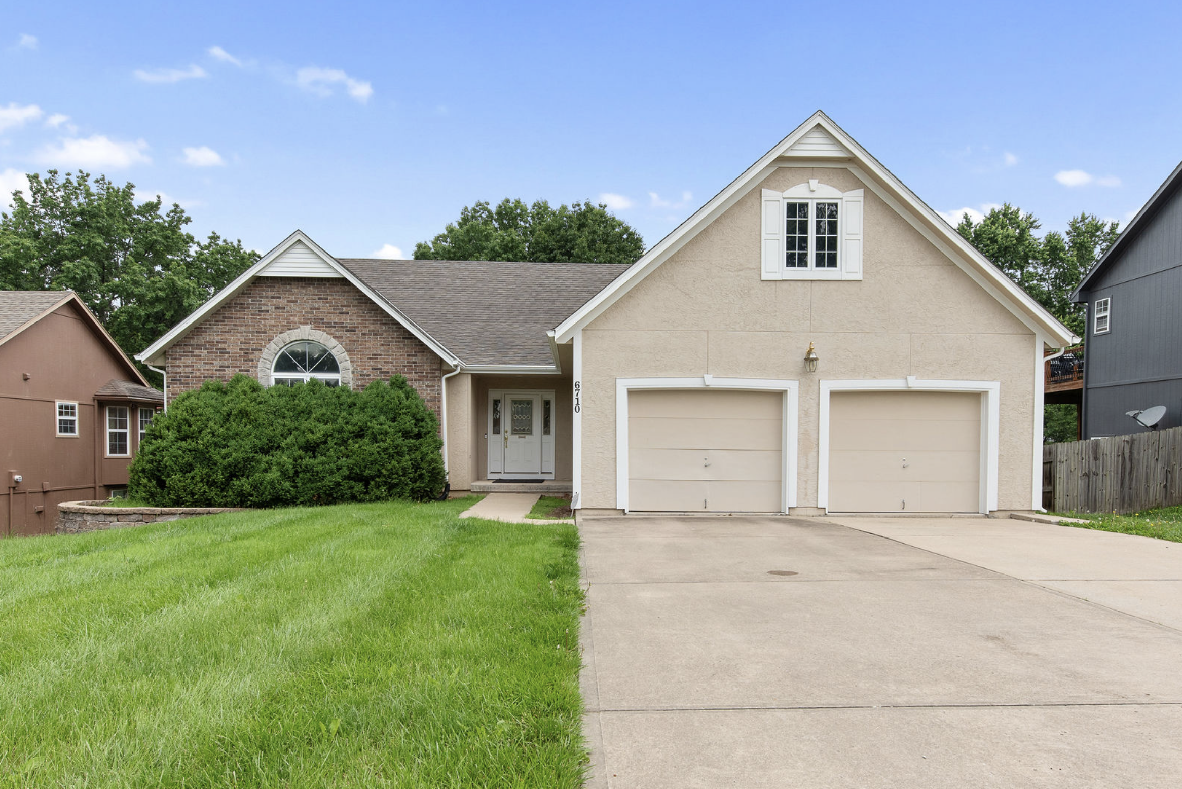 Front view of a suburban house with a garage, beige walls, white trim, and a brick section, surrounded by green lawn and trees under a blue sky.