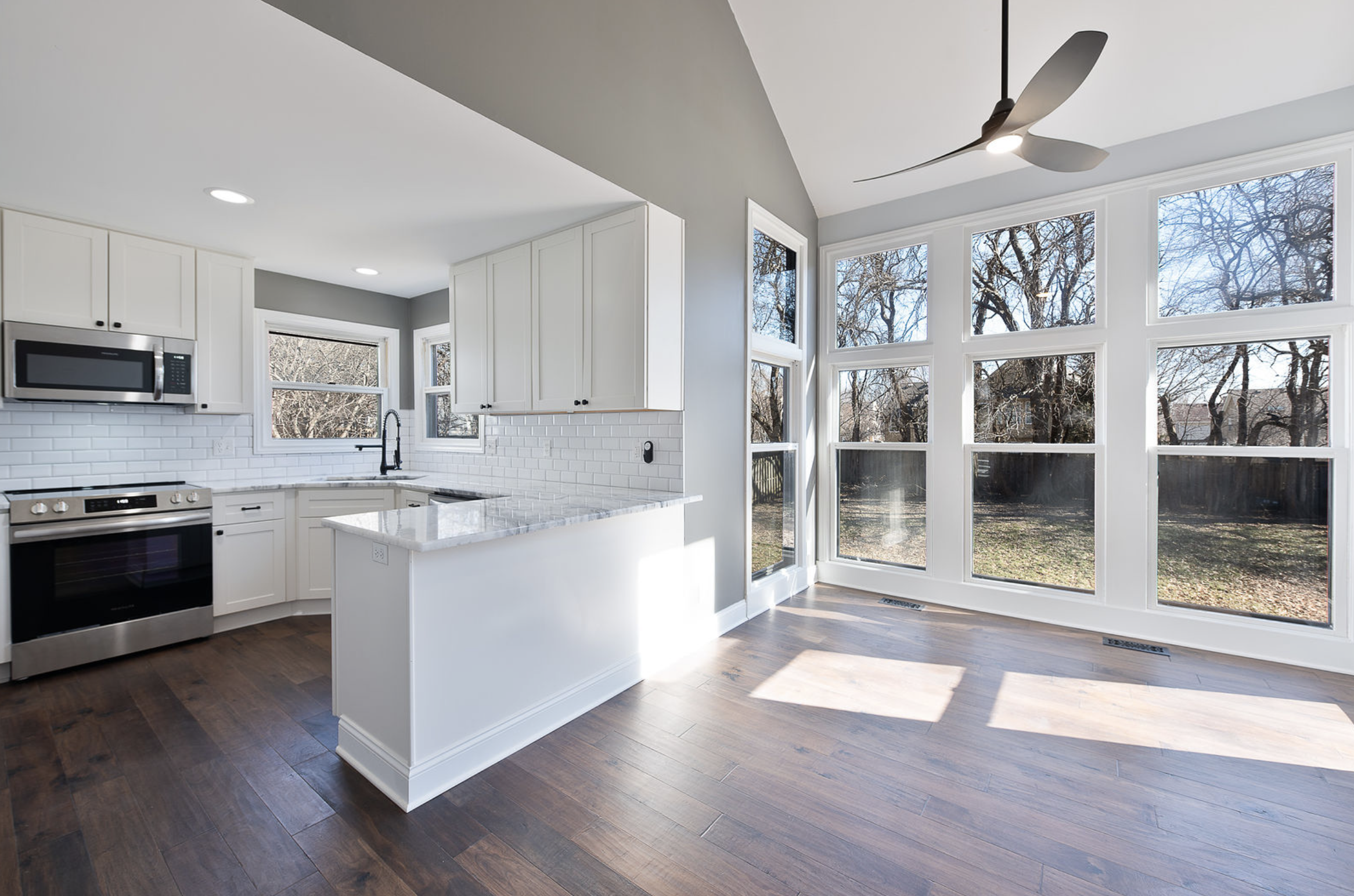 Kitchen with white cabinets, stainless steel appliances, a breakfast bar, large windows with a view of the backyard, and a ceiling fan.