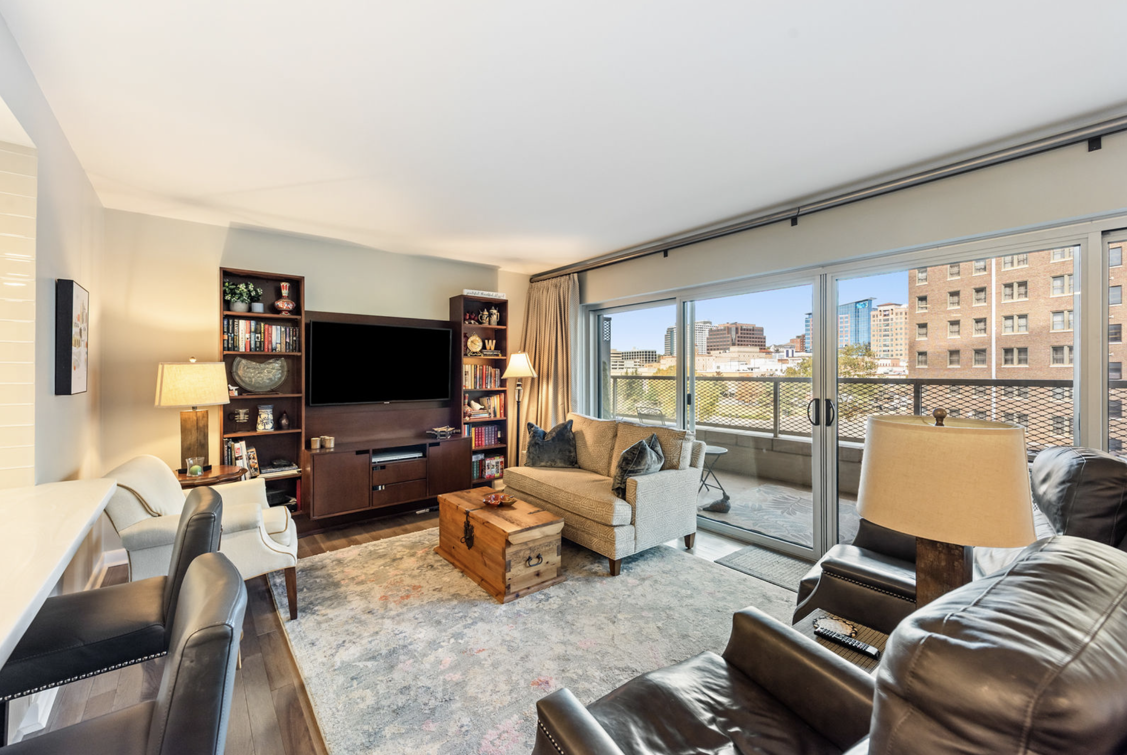Living room with balcony view, sofa, armchairs, wooden coffee table, TV, bookshelf, and decorative lamps.