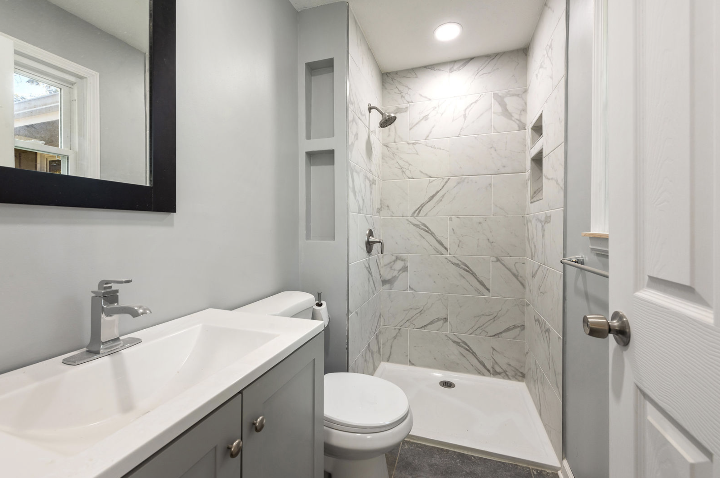 A modern bathroom with a white vanity with a sink and faucet, a large mirror, a toilet, and a walk-in shower with marble tile walls and built-in shelves.