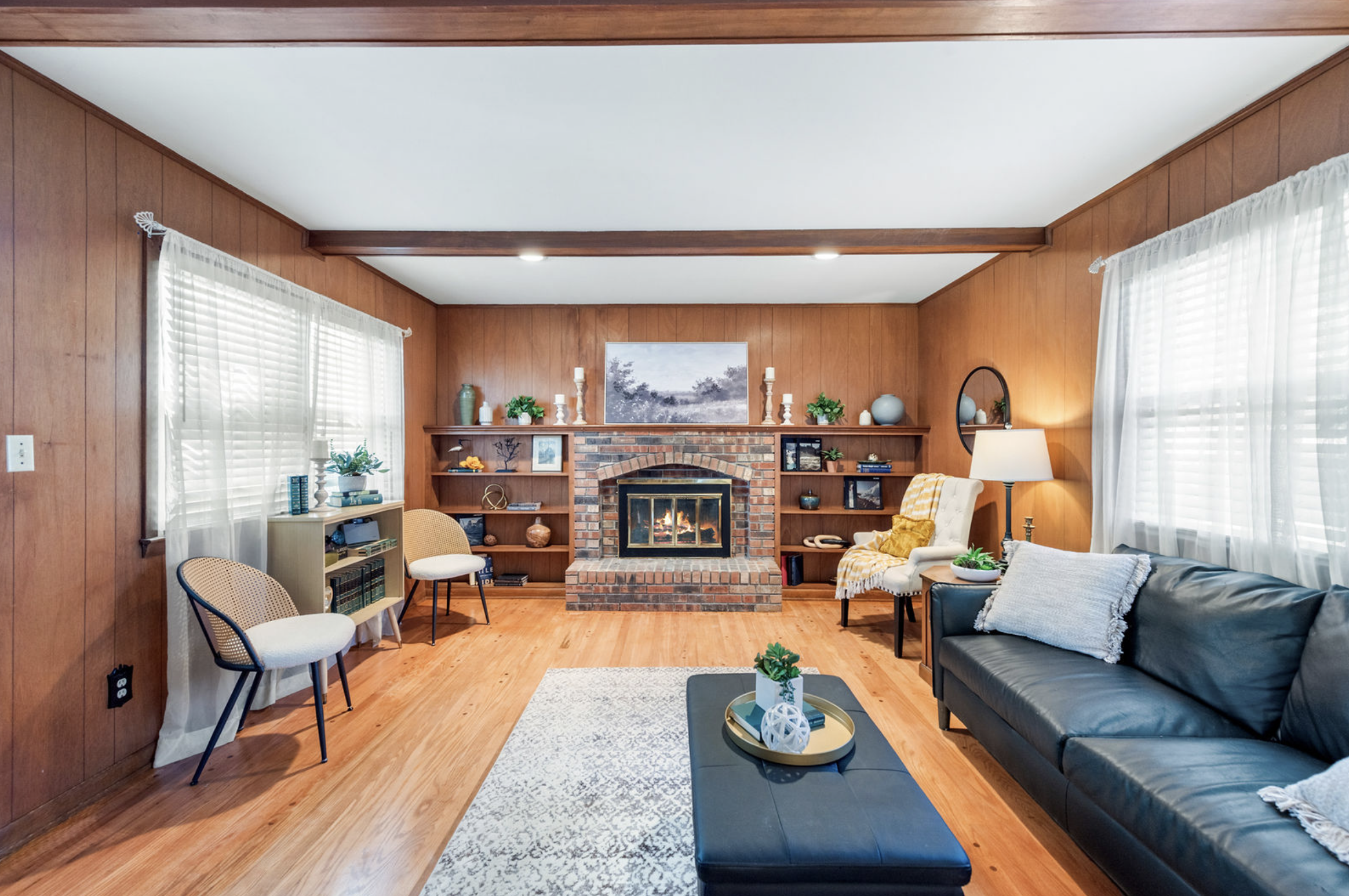 Living room with wood-paneled walls, large windows with white curtains, a brick fireplace, and modern furniture including a black leather sofa, beige chairs, a white armchair, and decorative items on shelves and tables.