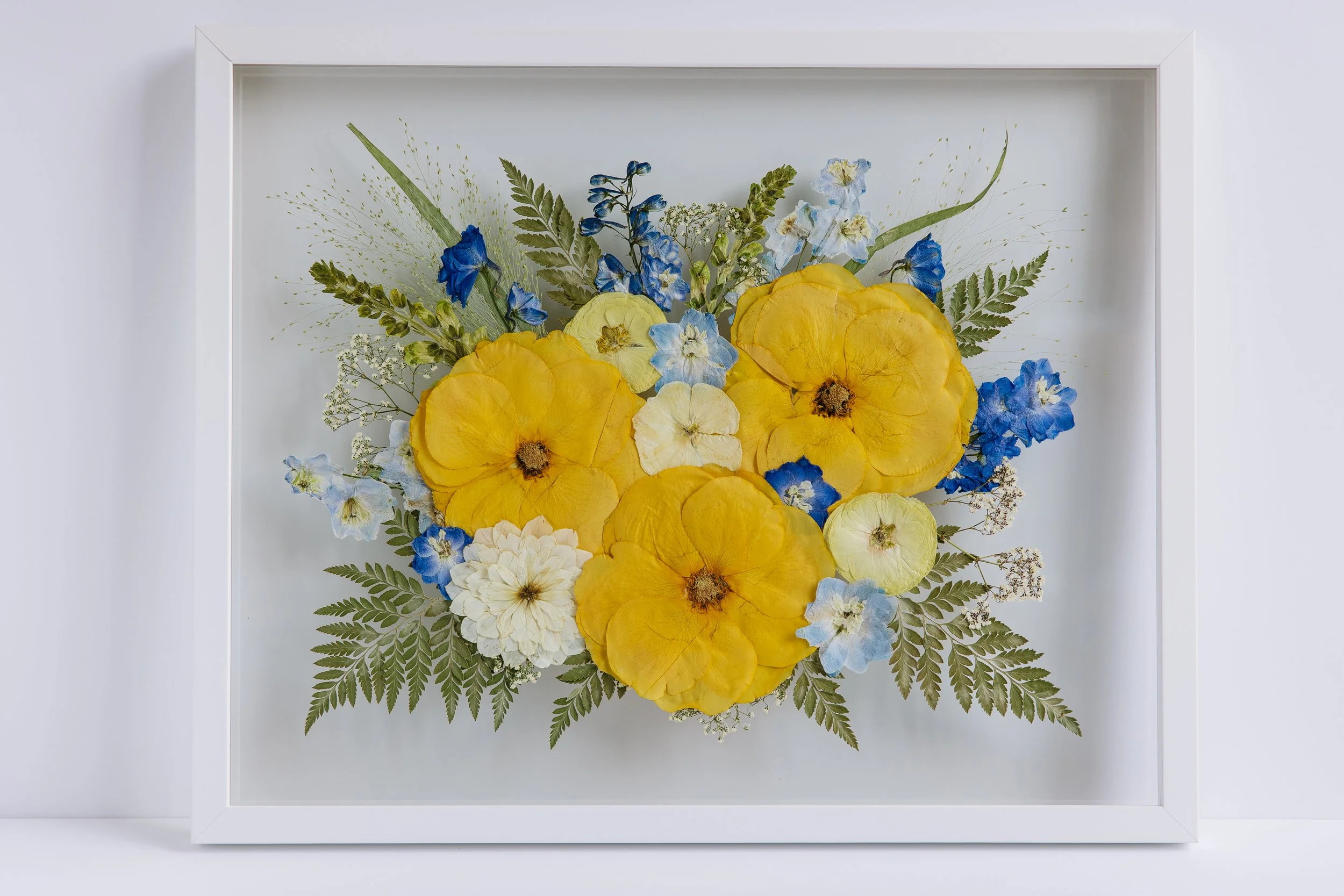A framed wedding invitation surrounded by pink, cream, and white flowers with green leaves, displayed on a concrete floor against a white wall.