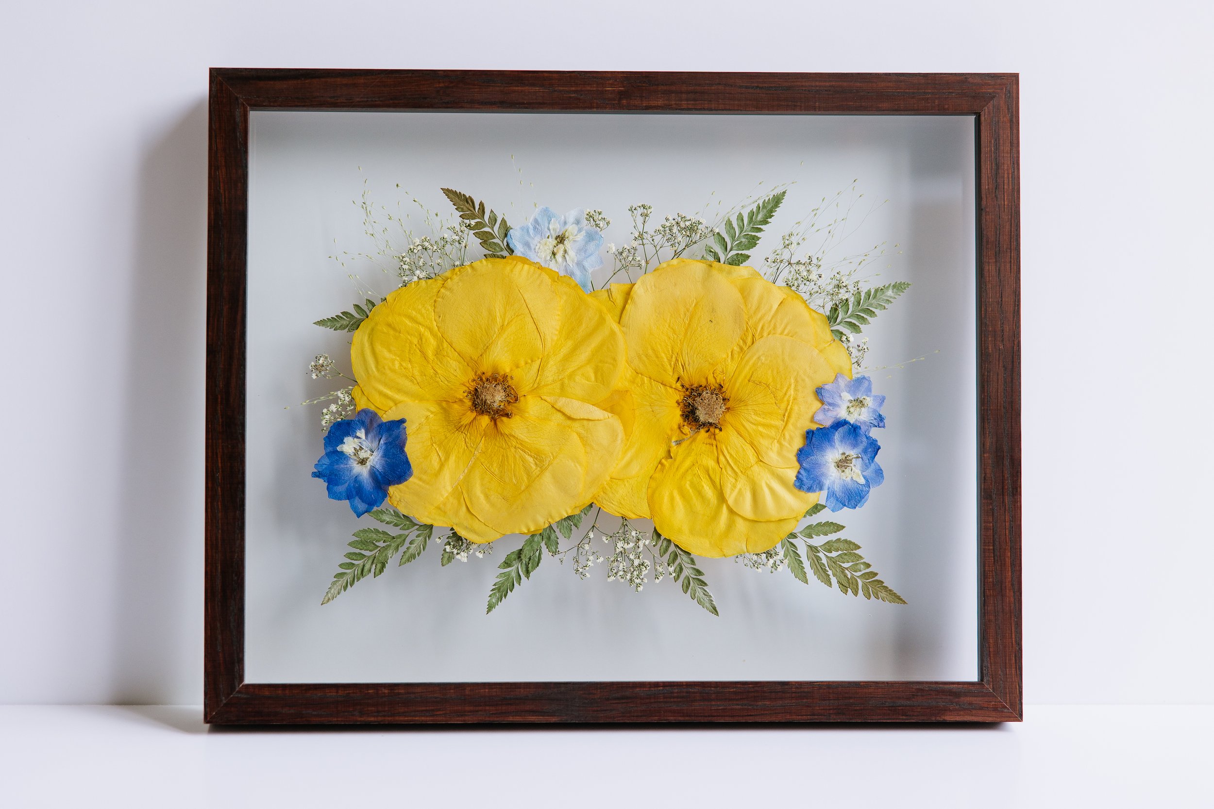A brown frame contains two yellow roses and other blue flowers.