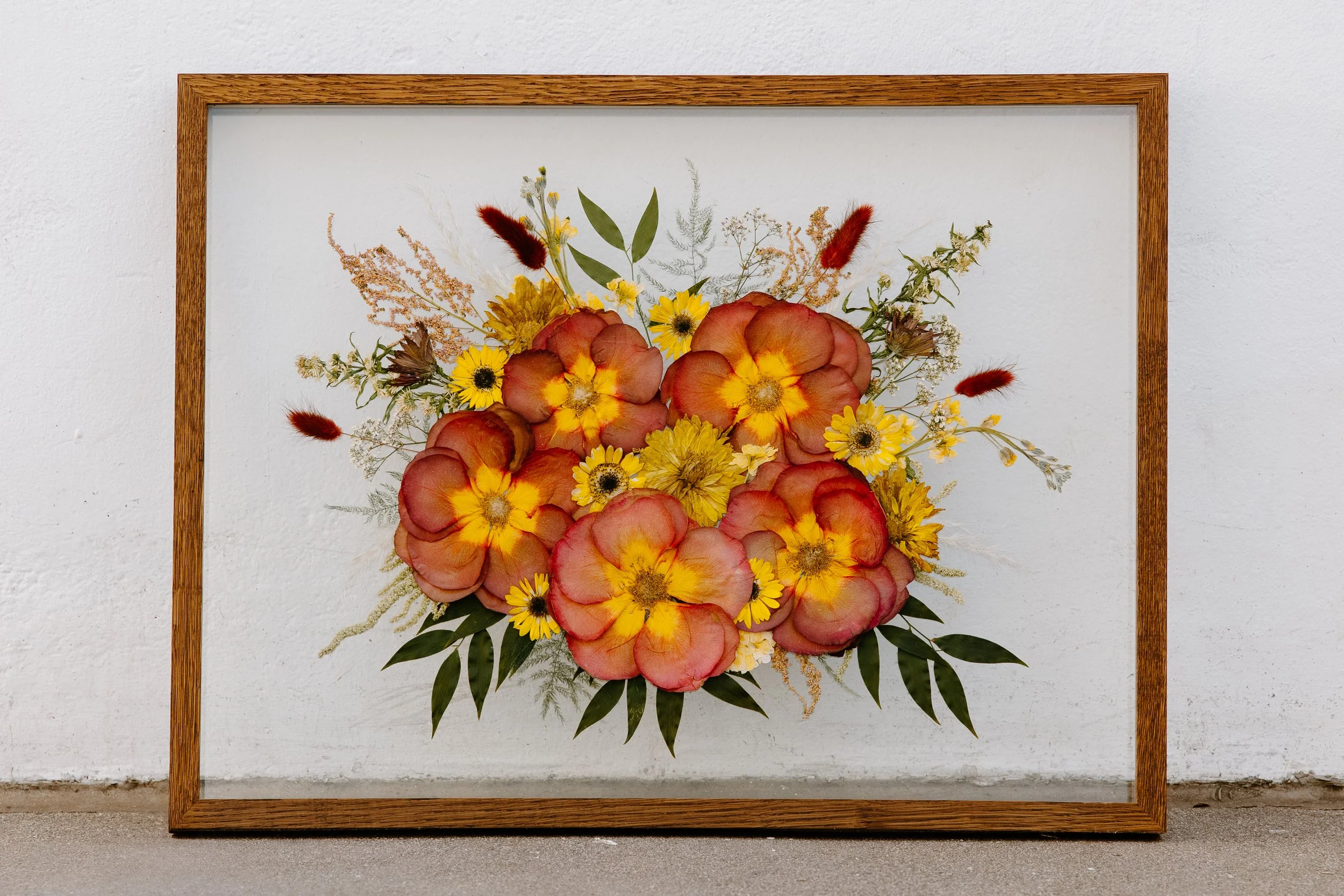 A pressed floral frame leans up against a white wall. The frame is full of red, orange and yellow flowers.