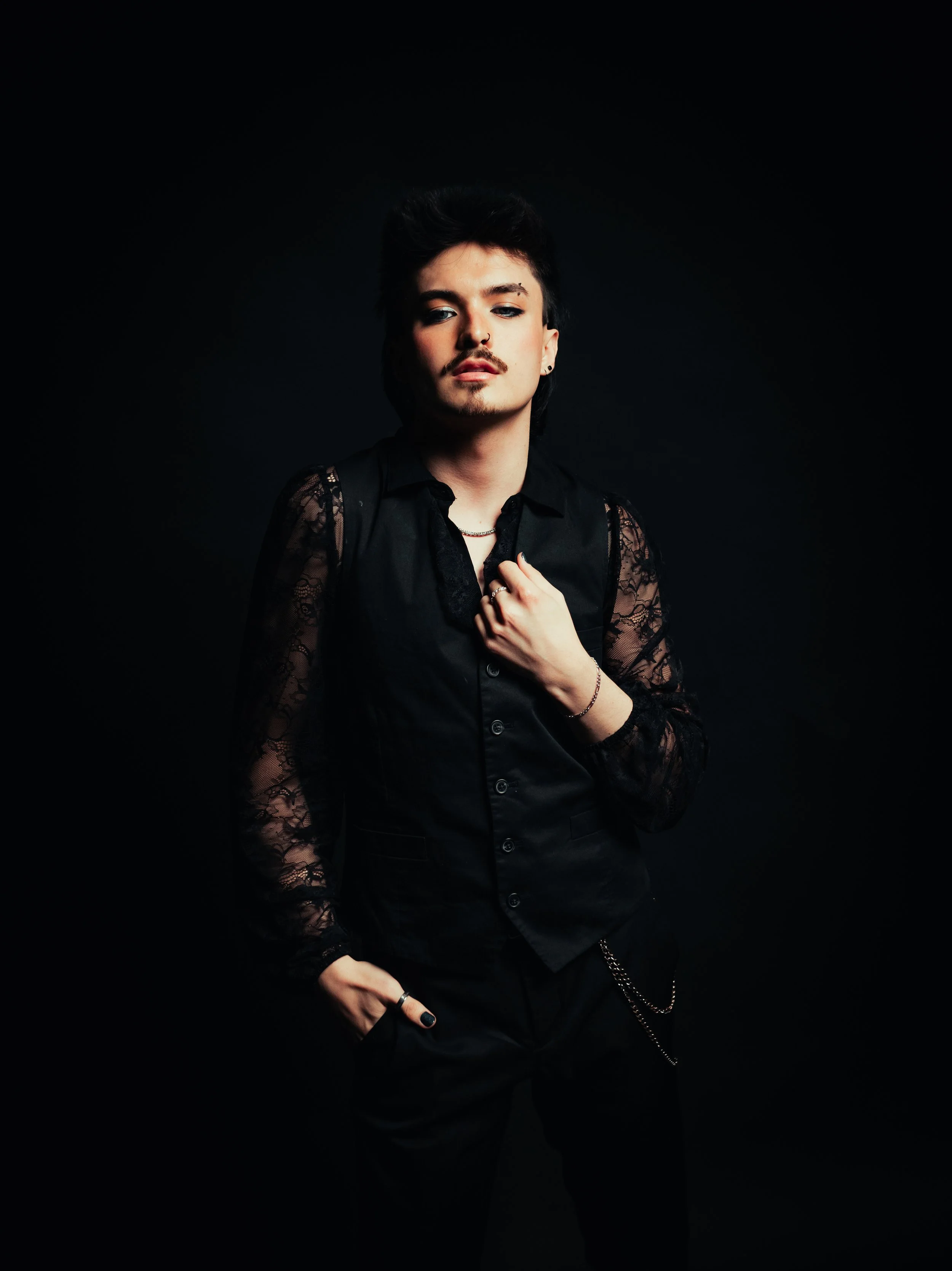 Person with short dark hair, wearing a black leather jacket, fishnet shirt, and black pants, standing against a red background.