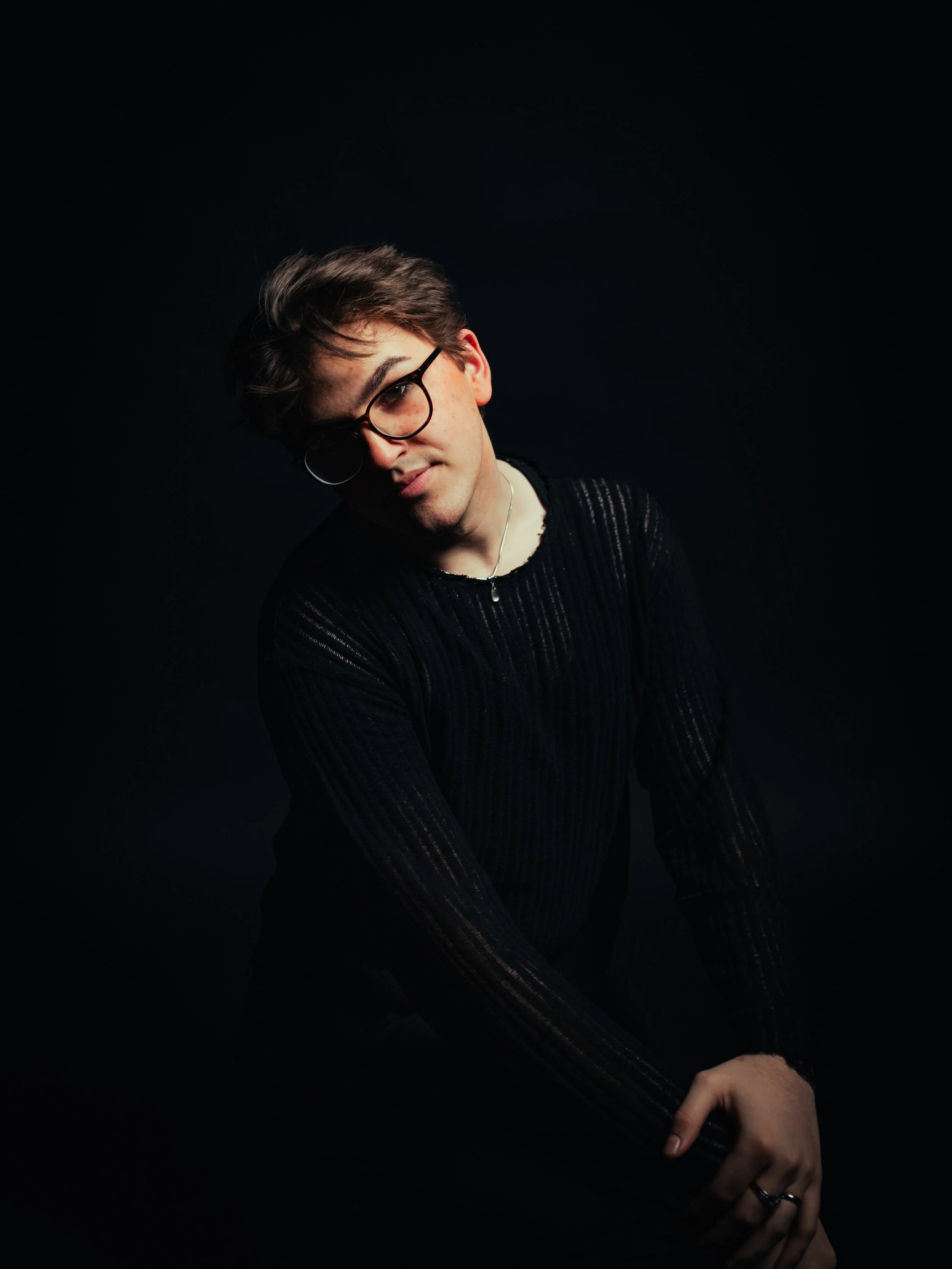 A person with short dark hair, glasses, and jewelry, wearing a black lace shirt, standing against a red background, with one arm raised behind their head and a relaxed expression.