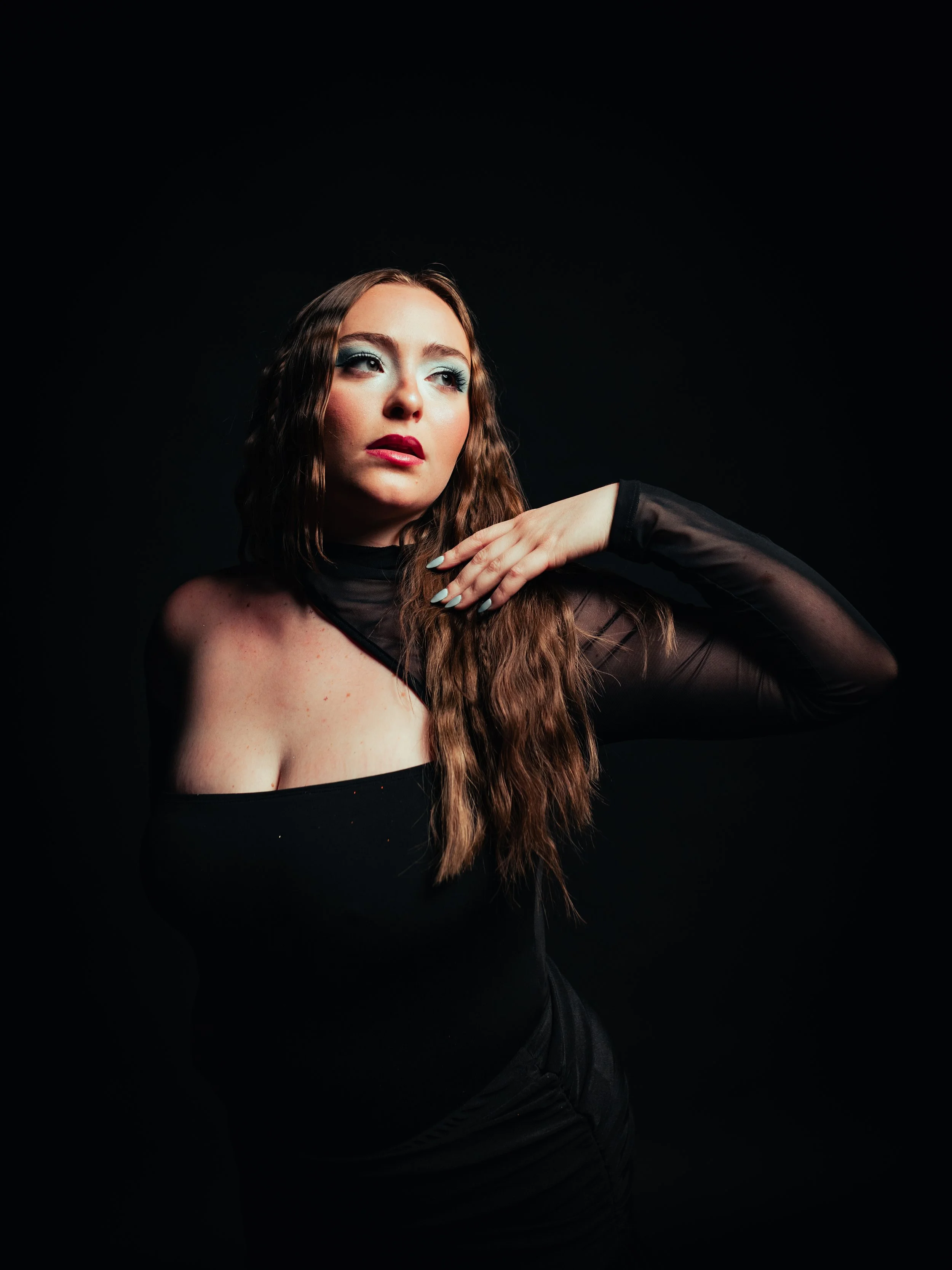 A young woman with long wavy brown hair wearing a fitted black dress with a feather detailing neckline, standing against a solid red background.
