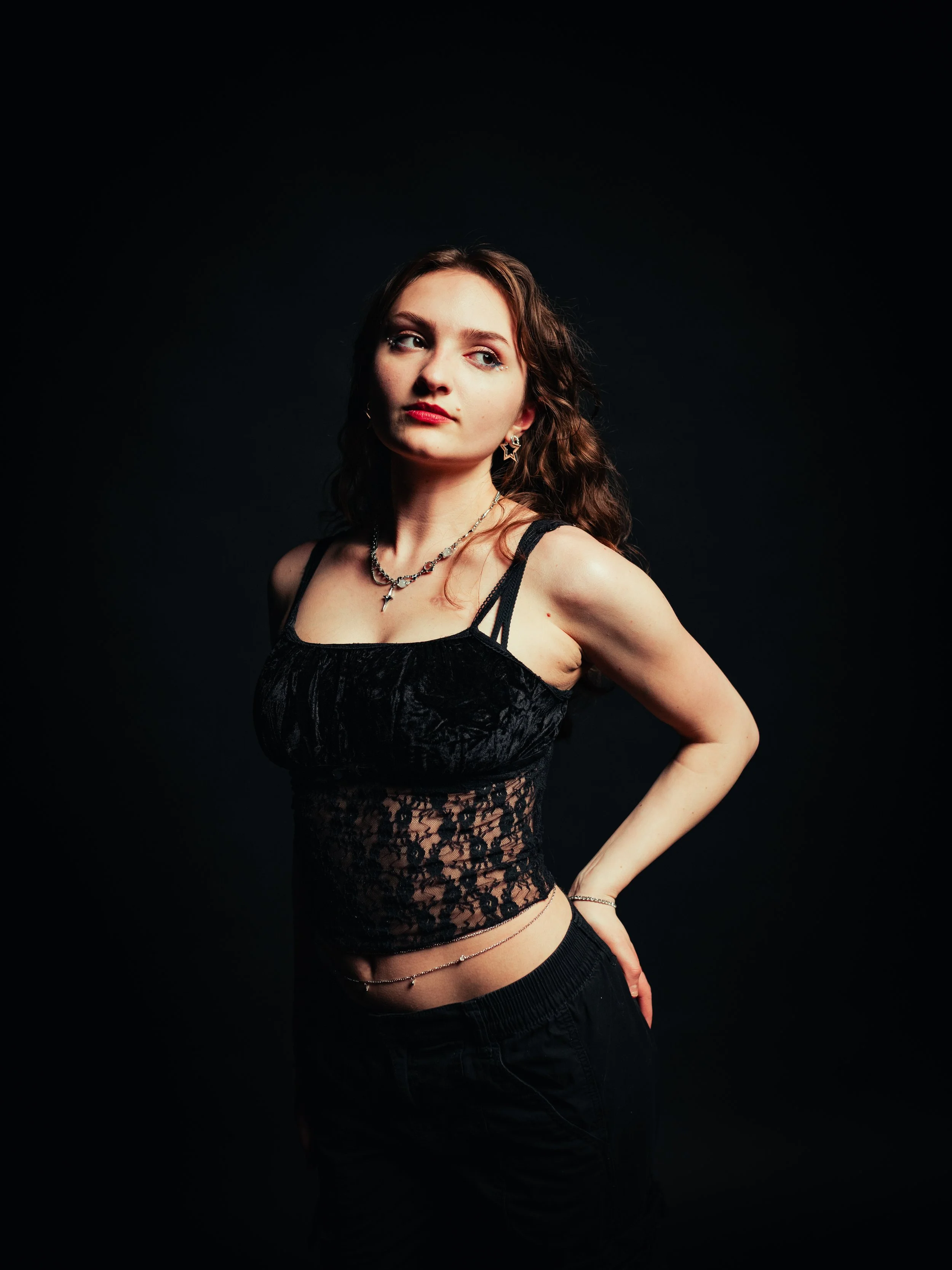 A young woman in a black dress and sheer tights standing with hands on hips against a red background.