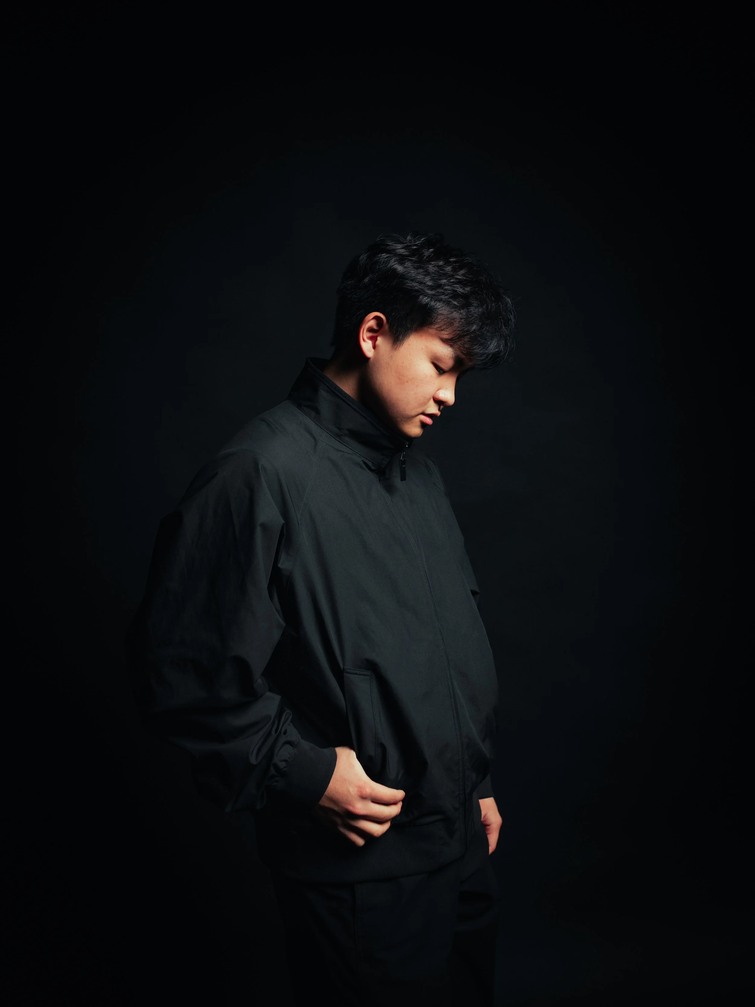 Young man with dark hair, wearing a black leather jacket and black pants, standing against a red background, with one hand behind his head, looking to the side.