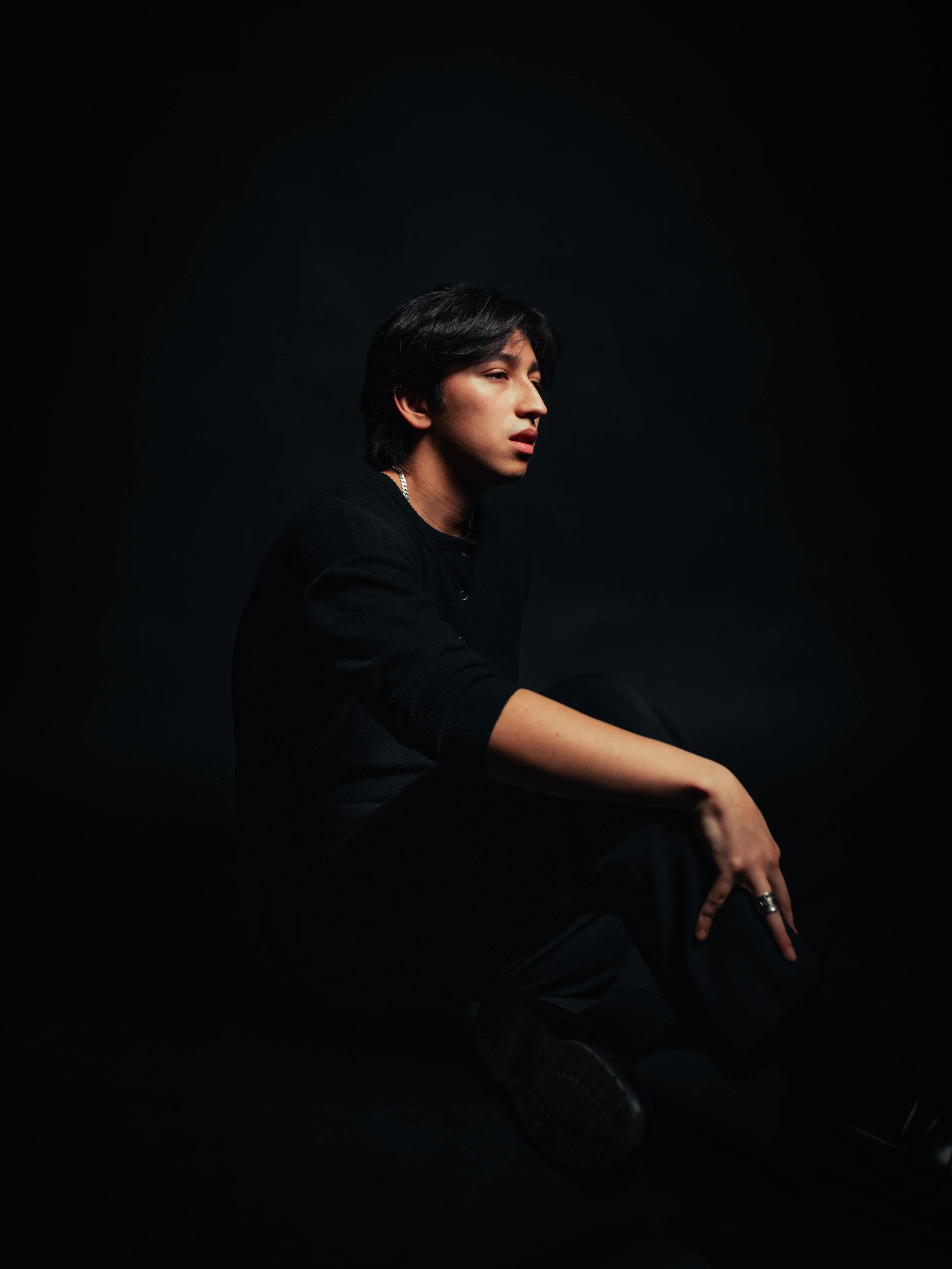 Young man with black hair in a black suit, posing against a red background, wearing jewelry including a necklace and bracelet.