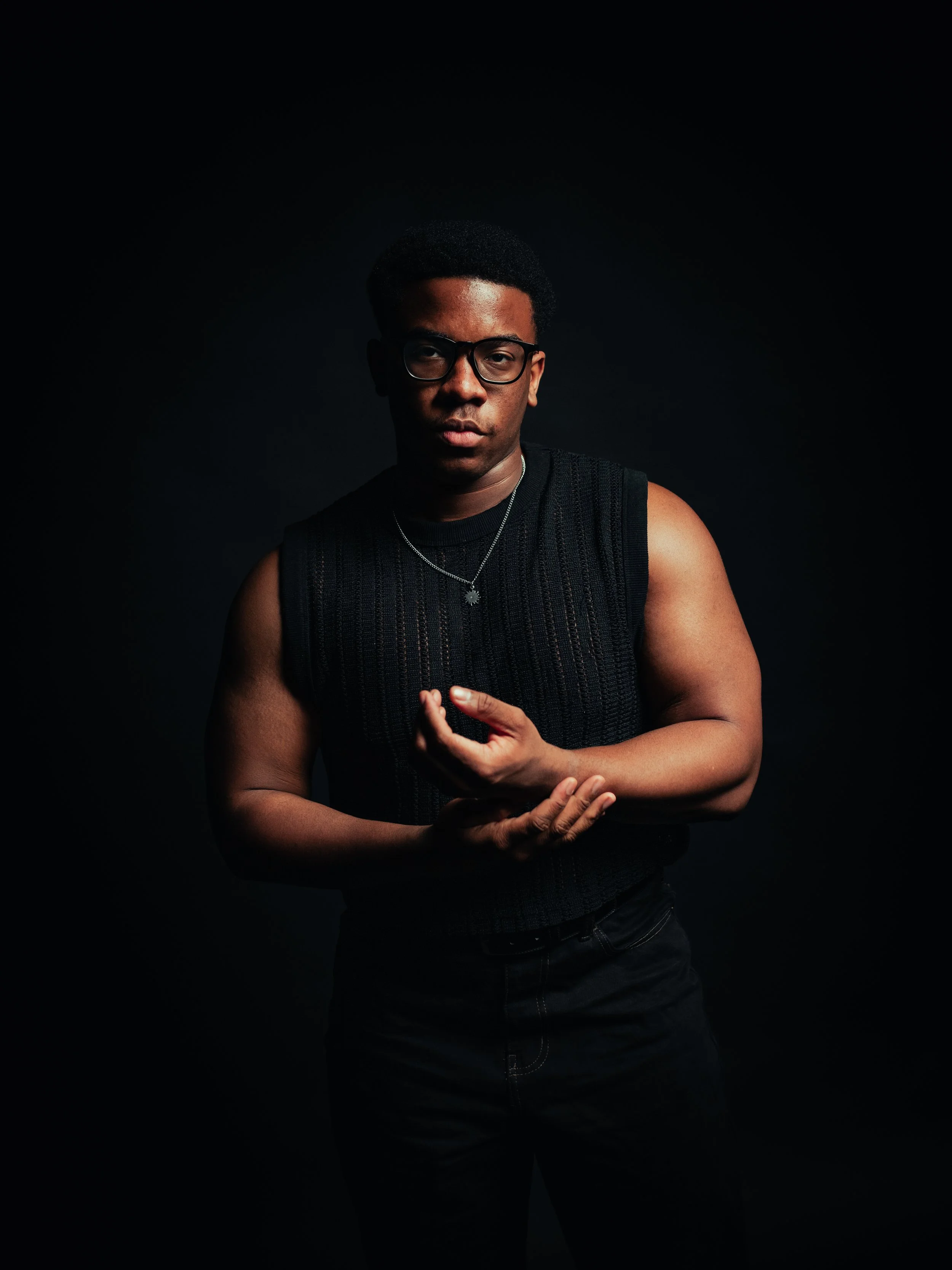 A young man wearing glasses, a sleeveless black shirt, and a chain necklace, standing against a red background with one hand over his chest and the other in his pocket.