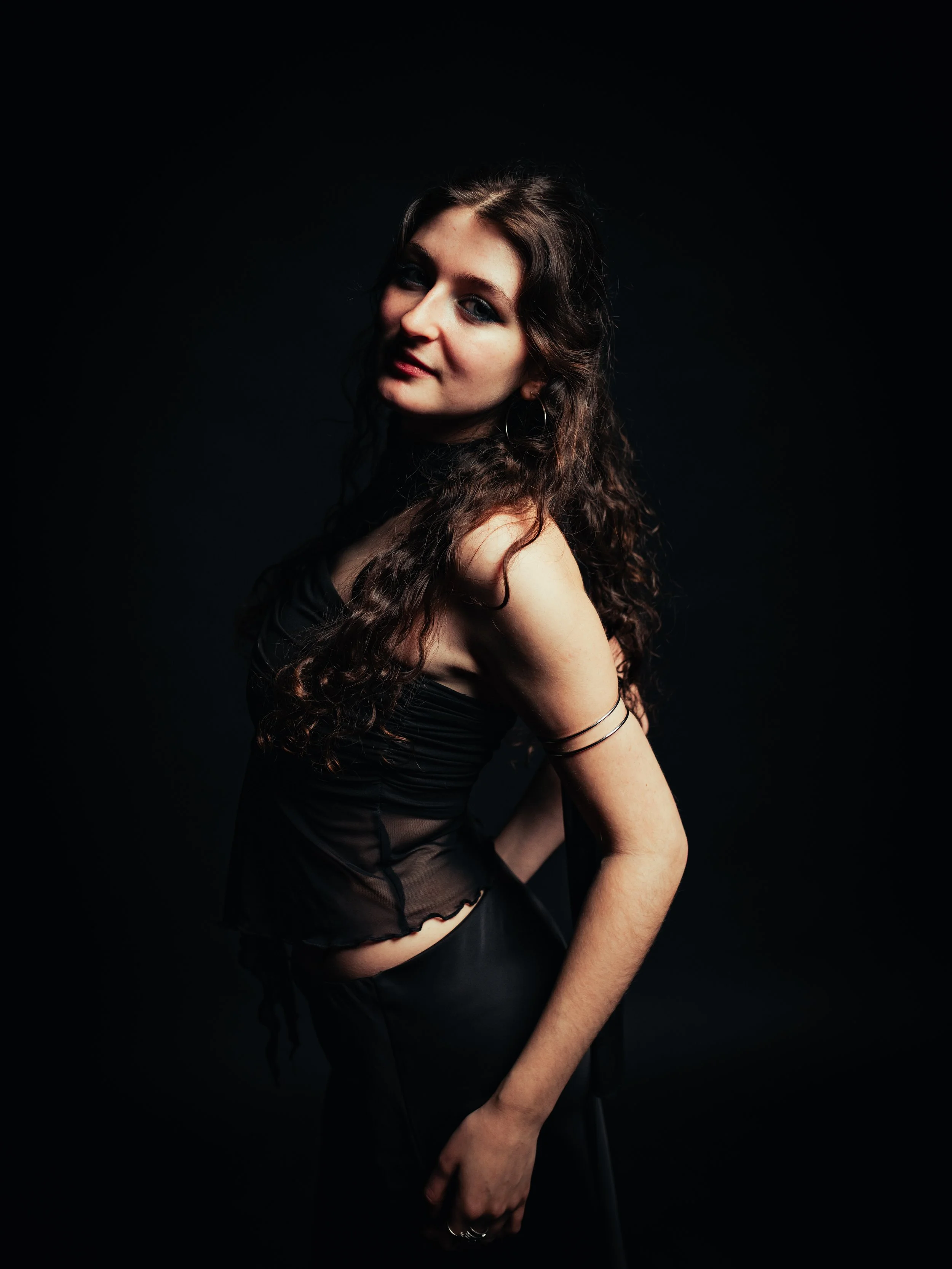 A young woman with long brown hair and blue eyes stands against a red background, wearing a black sleeveless top, a black skirt, and black tights.