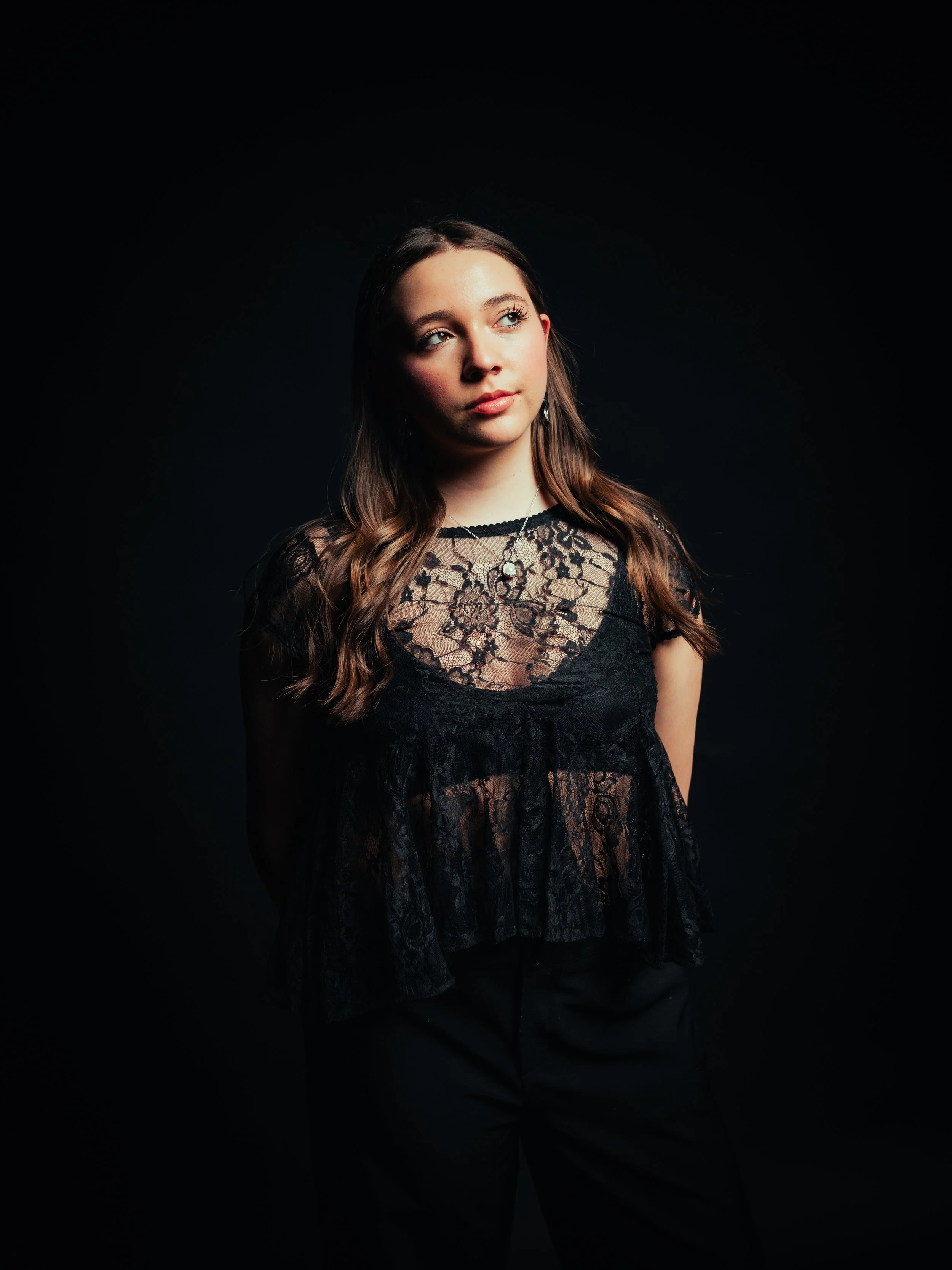 Young woman with long wavy brown hair wearing a sleeveless black dress, standing against a solid red background, looking slightly to the side with a neutral expression.