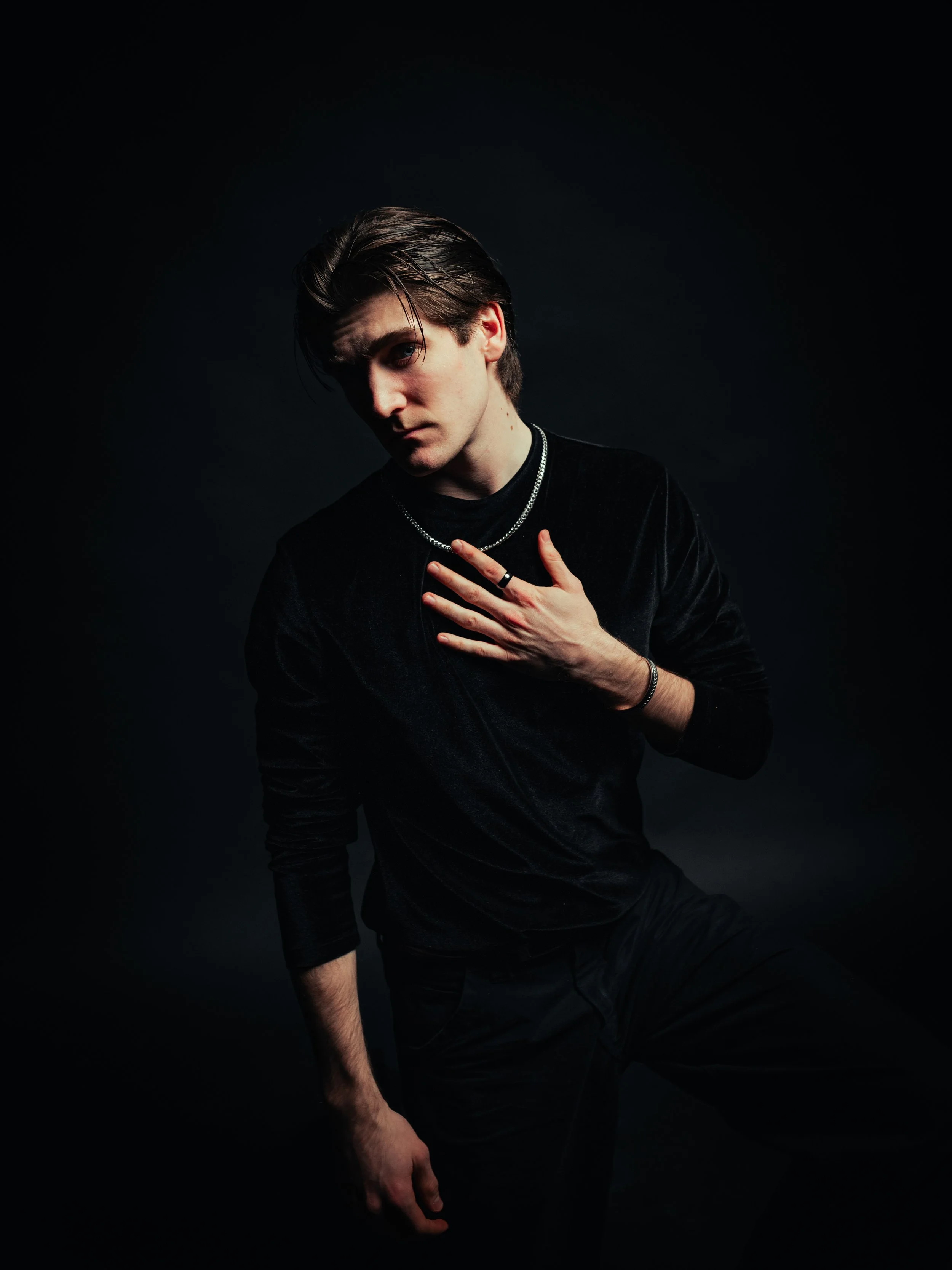 Young man with brown hair and fair skin wearing a black velvet long-sleeve shirt, silver jewelry, standing against a red background.