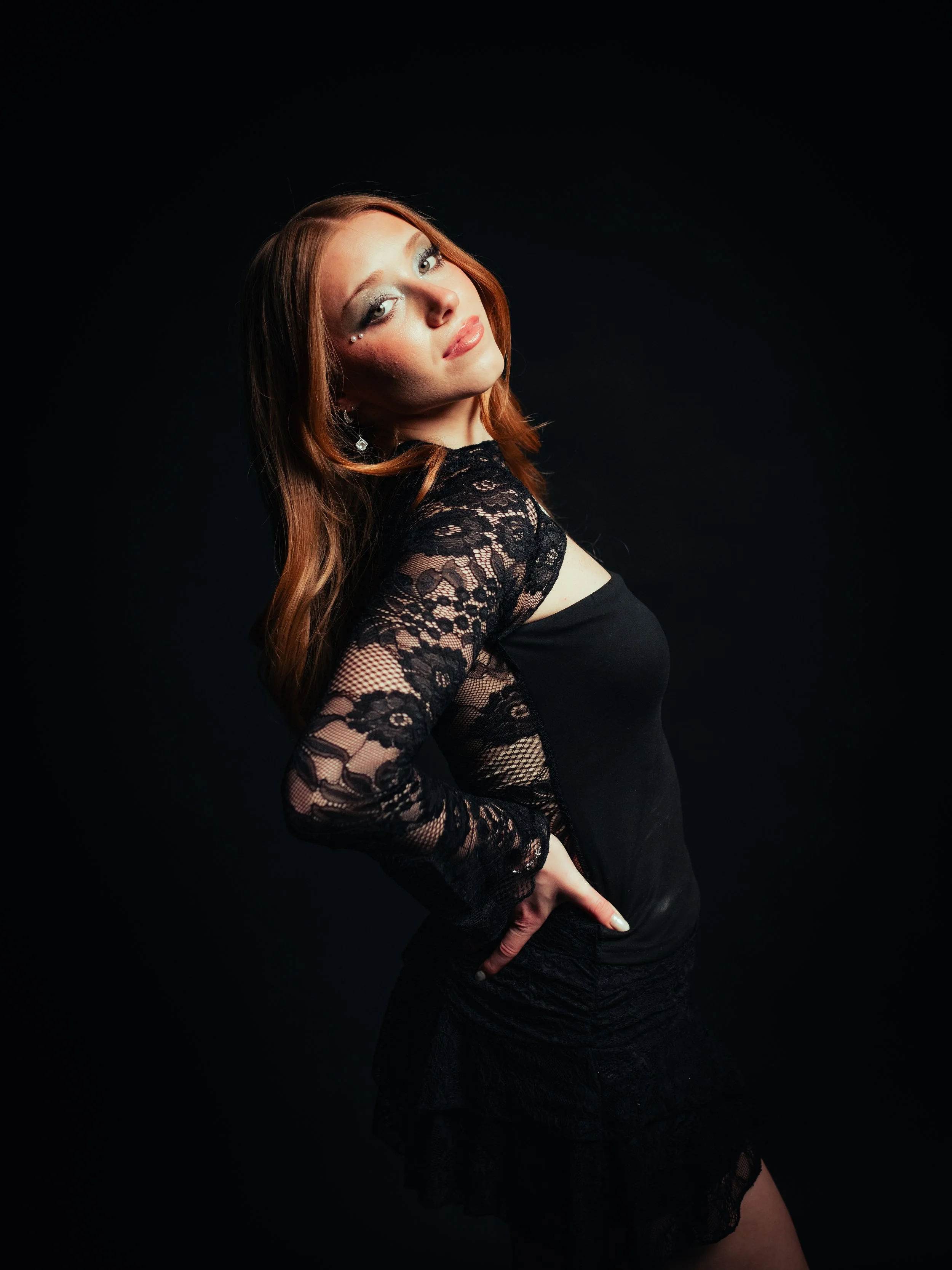Red-haired woman wearing a black lace top and black pants standing against a red background.
